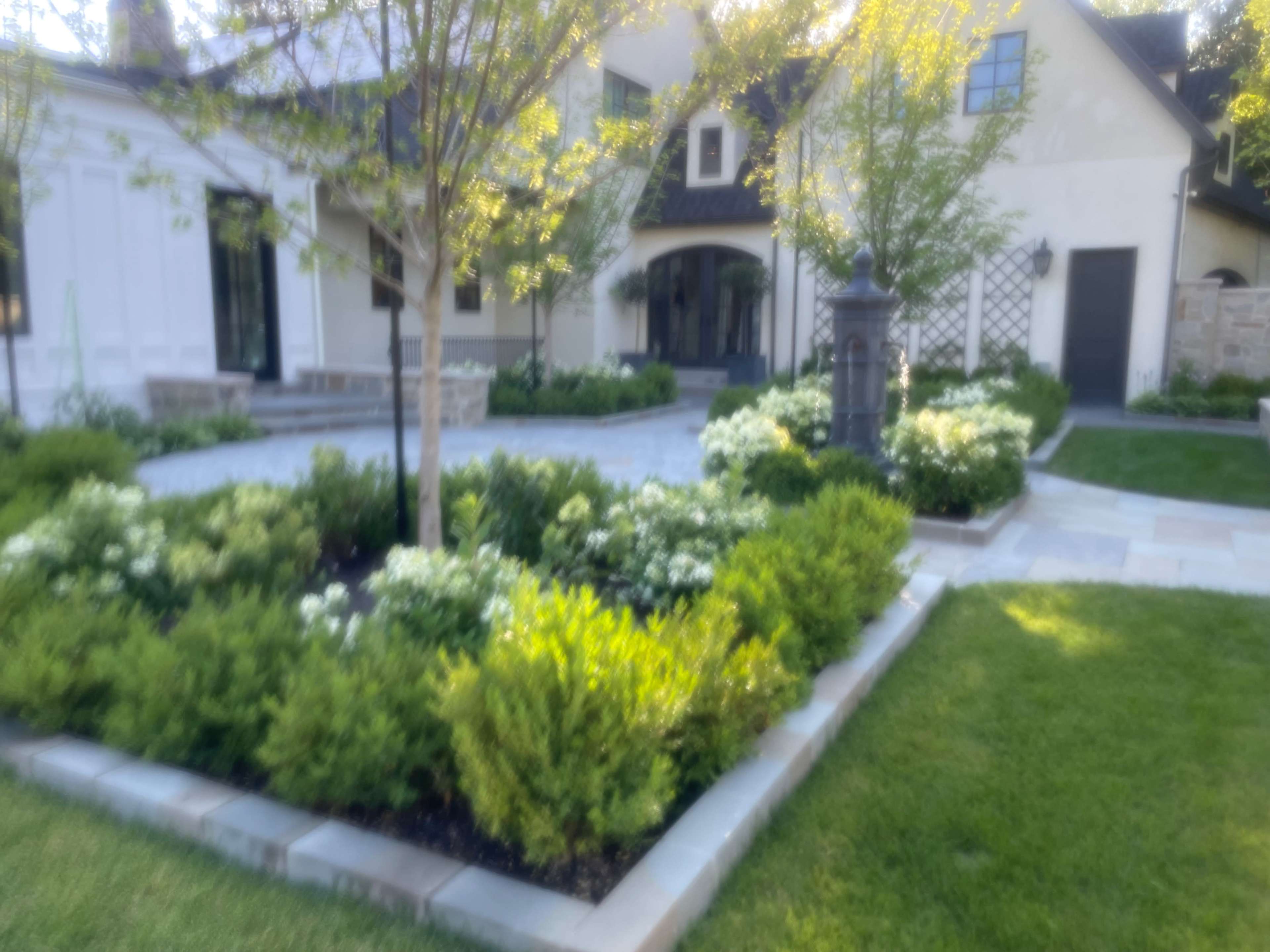 The image shows a landscaped garden featuring neatly trimmed bushes and flowering plants surrounding a stone patio in front of a modern house.