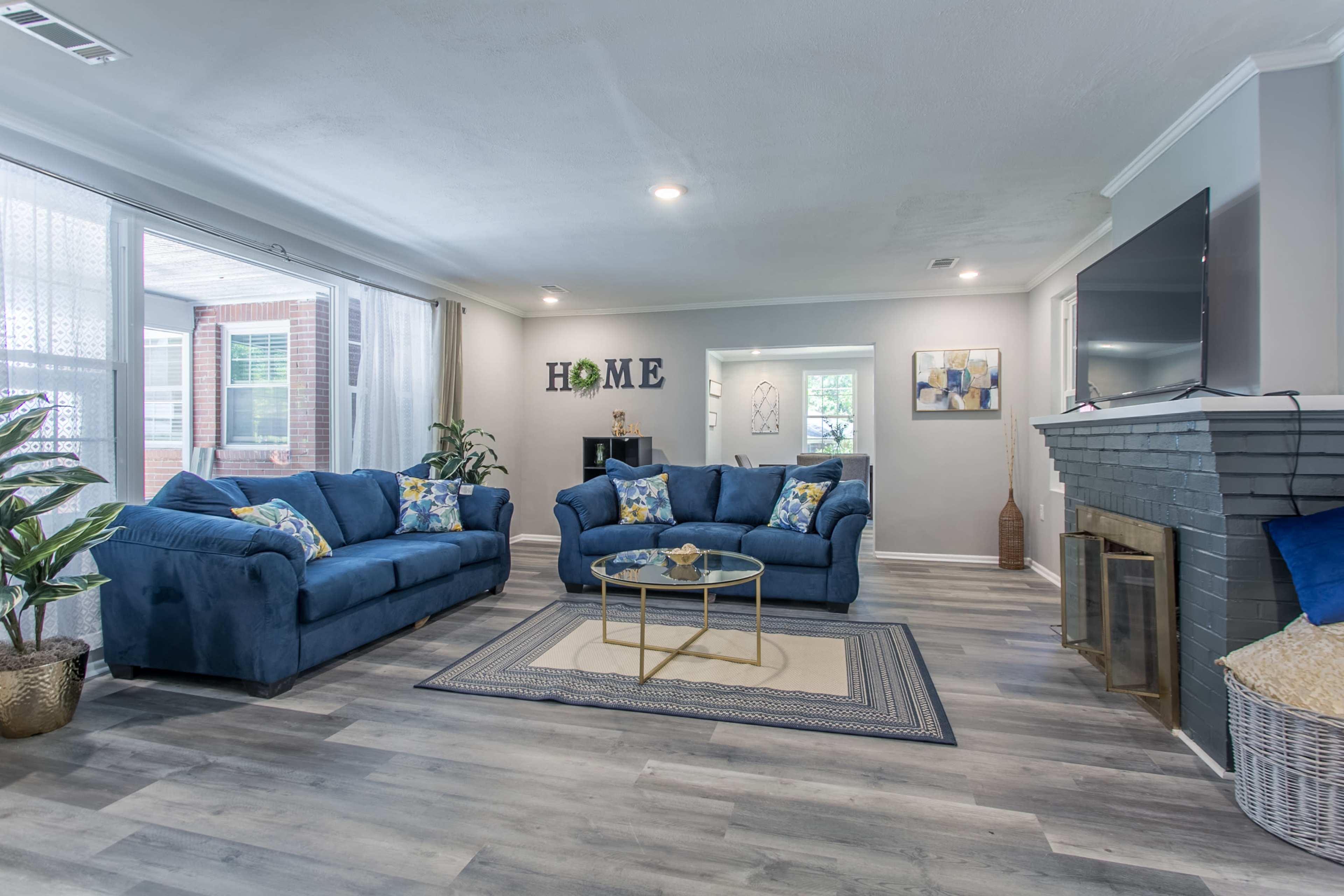 The living room features two blue sofas with decorative pillows, a glass coffee table, and a fireplace, with light streaming through large windows.