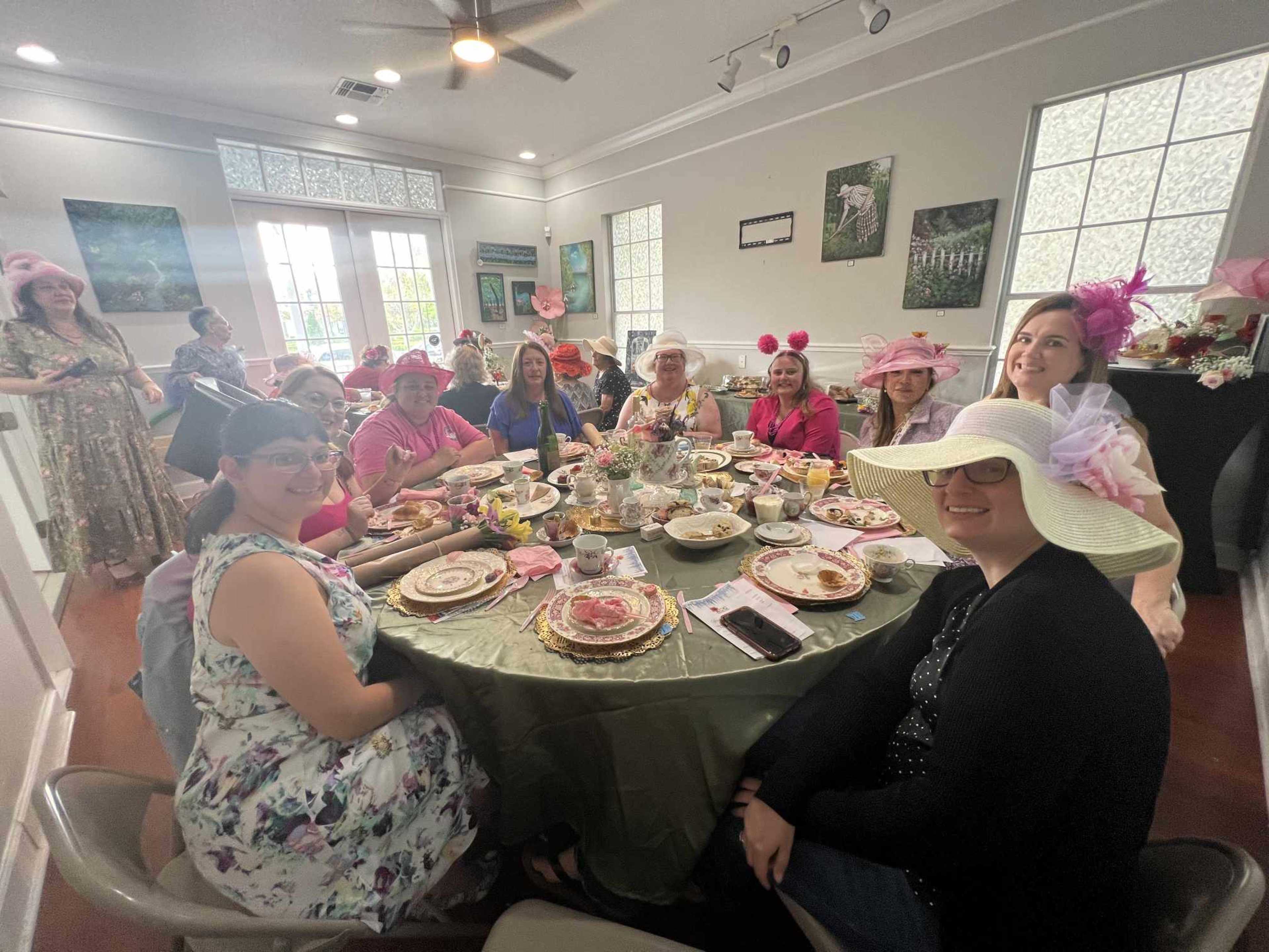 A group of women wearing large hats gathers around a table set for a tea party, with various dishes and decorative items in a well-lit room.