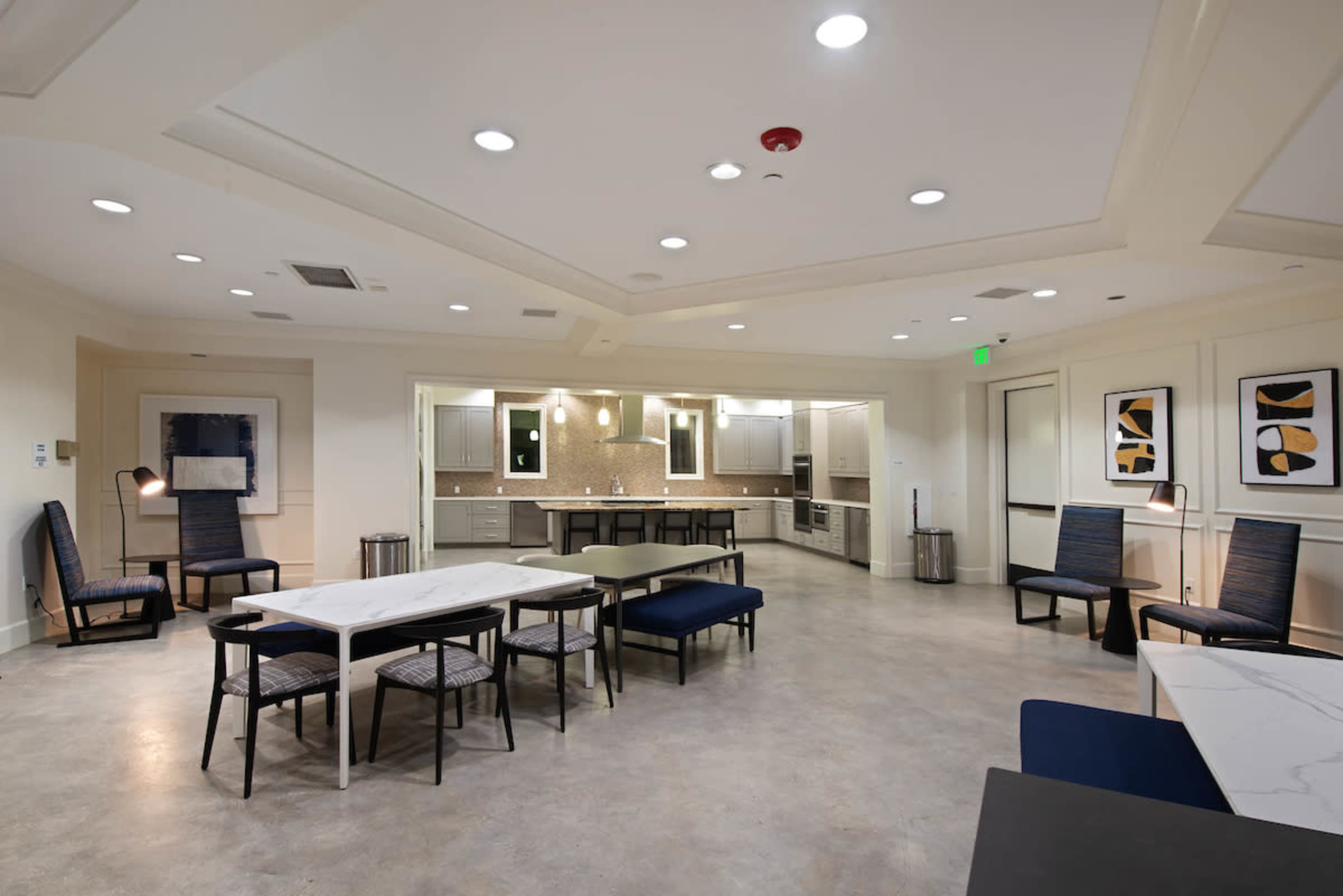 A spacious common area featuring tables and chairs, with a kitchen area in the background and contemporary lighting.
