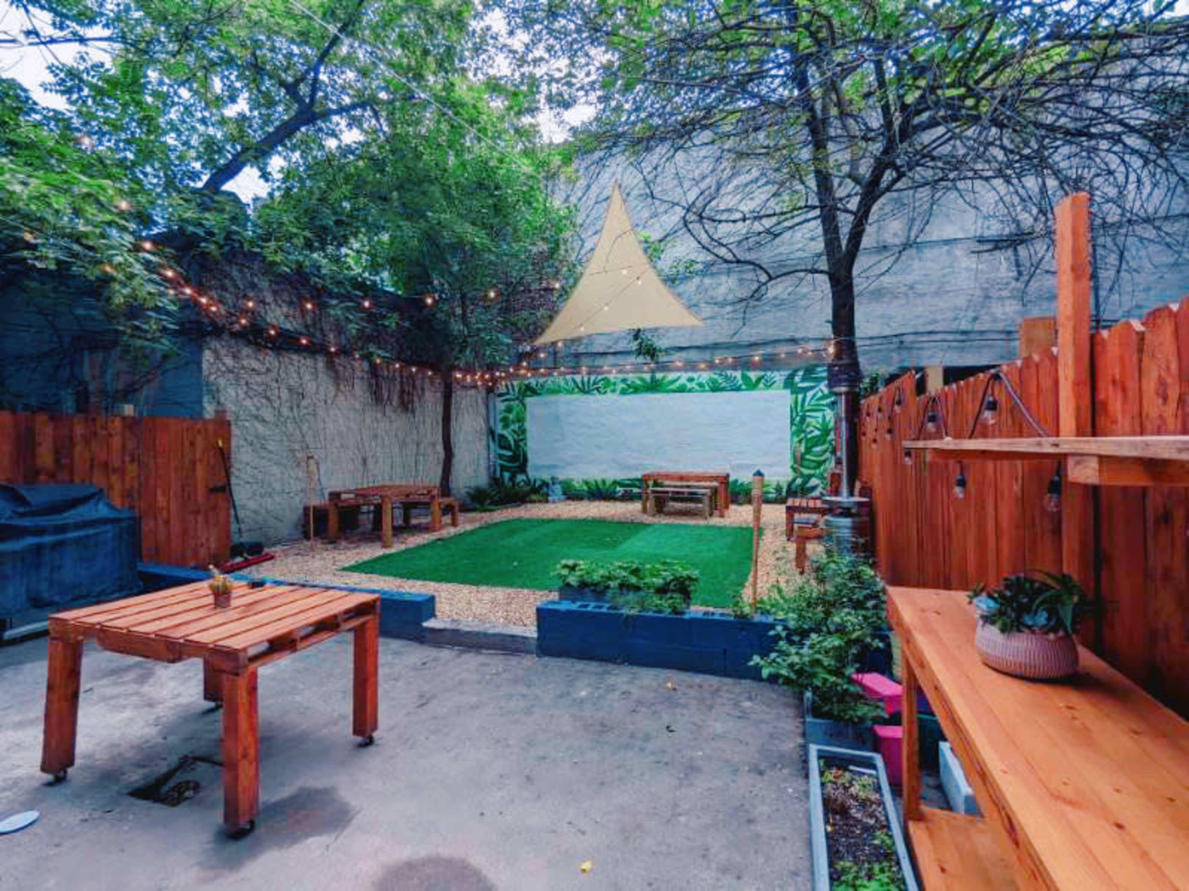 A backyard space featuring wooden benches, a grassy area, and a large white wall surrounded by greenery and string lights.