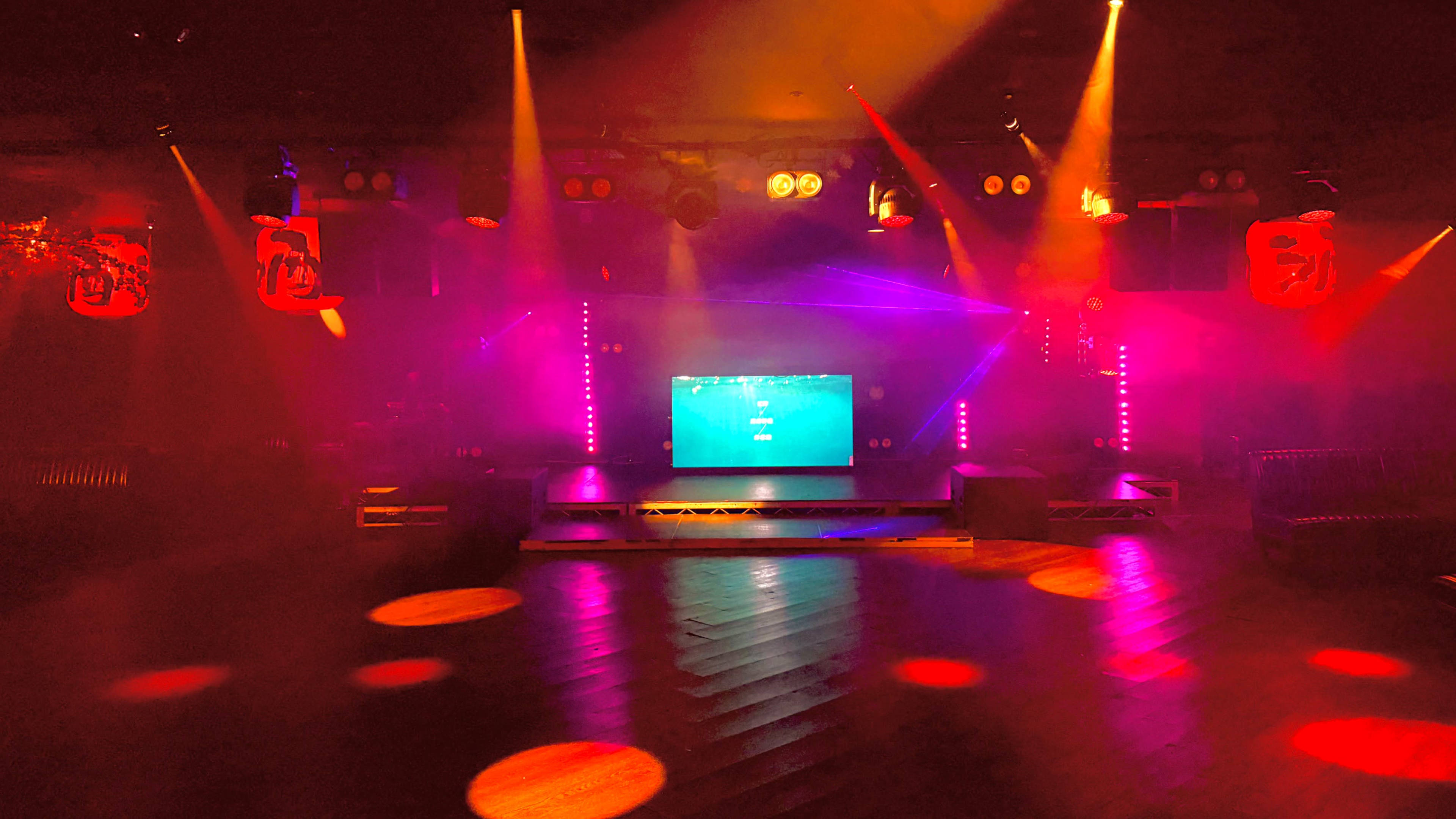 The image shows a dimly lit stage with colorful lighting effects and a central screen, set in a venue prepared for a performance or event.