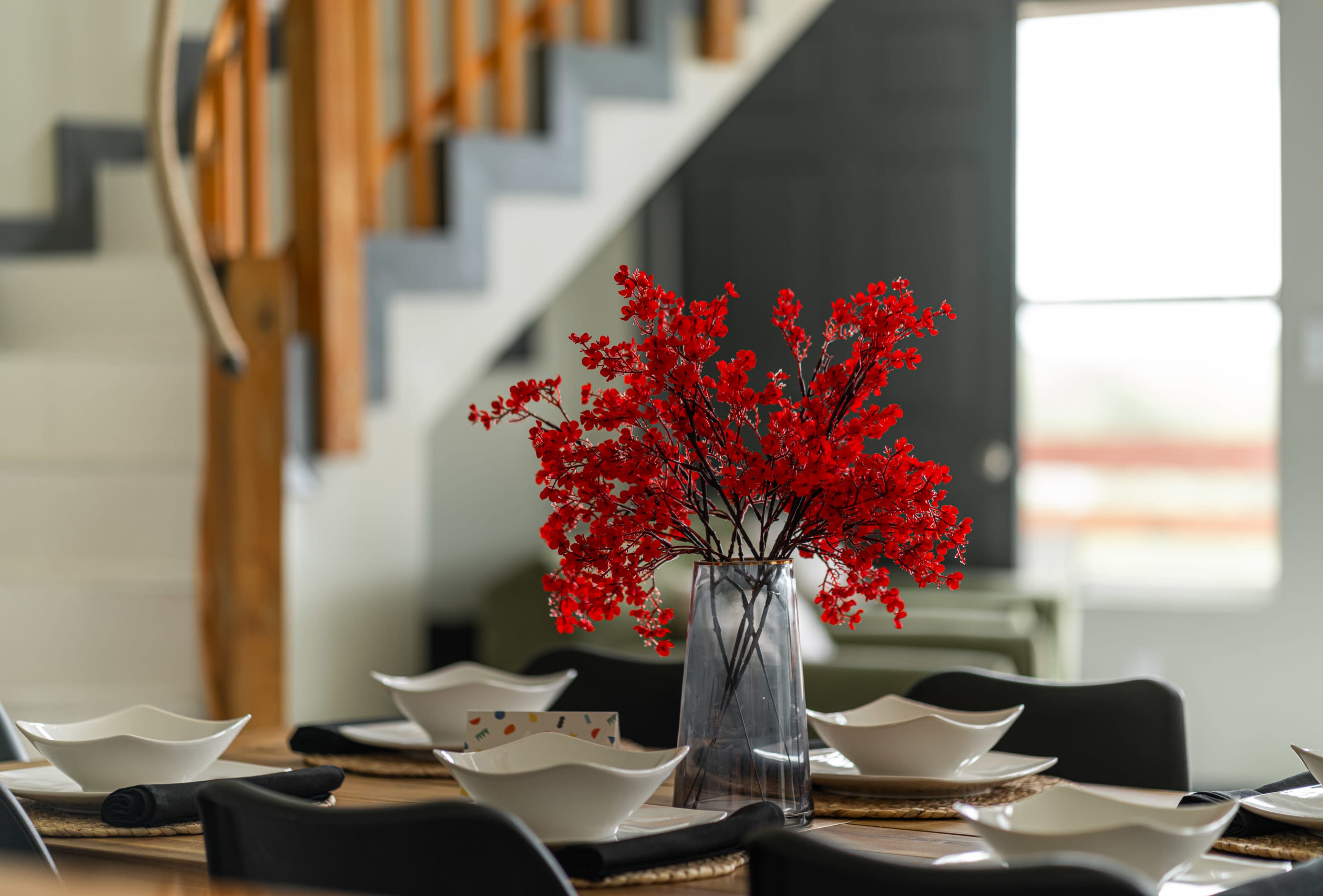 A table is set with white dishes and a vase of red flowers, with stairs and a door visible in the background.