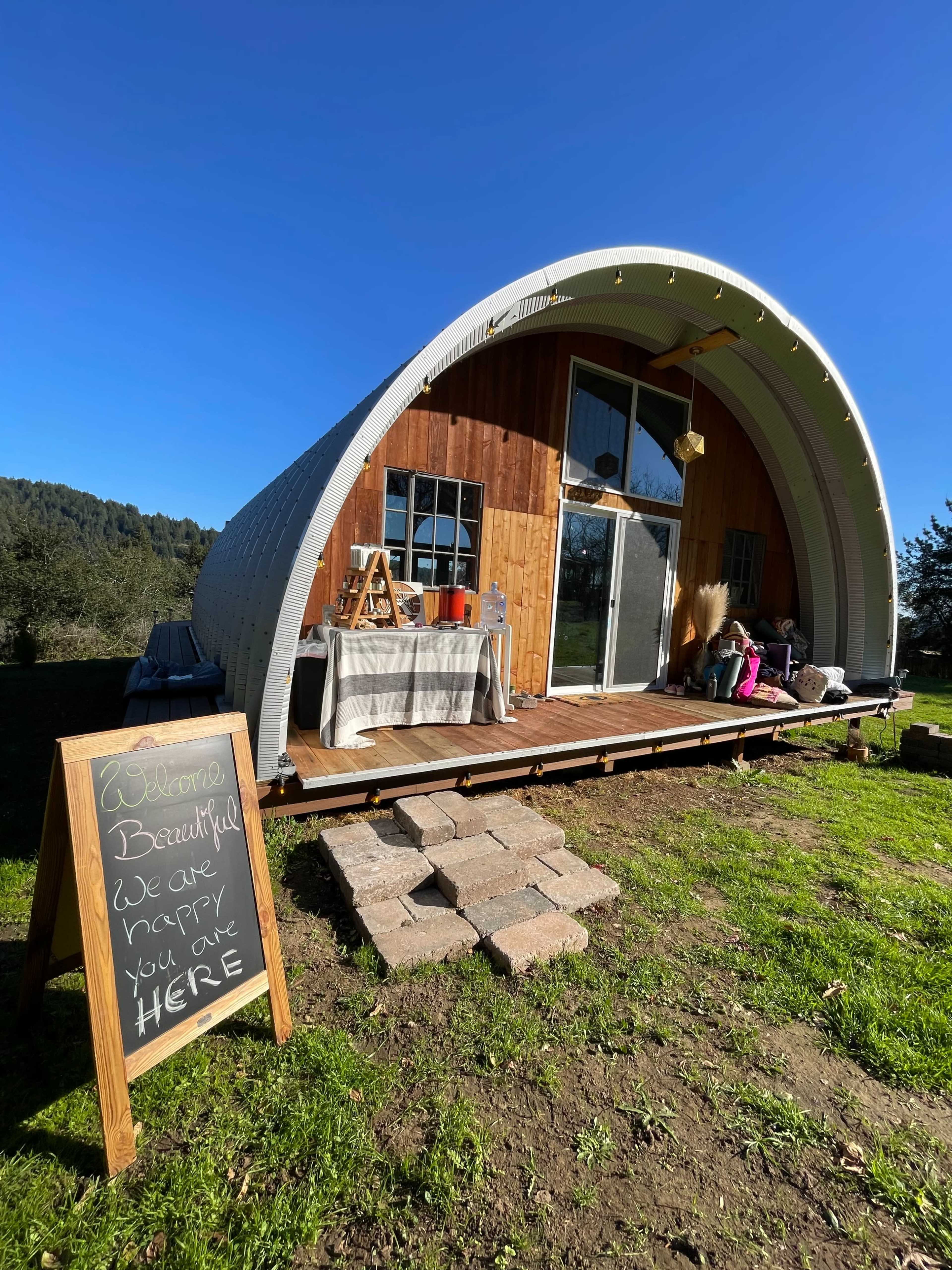 A curved, modern cabin made of wood and metal sits on a grassy area, with a welcoming sign next to steps leading to the entrance.