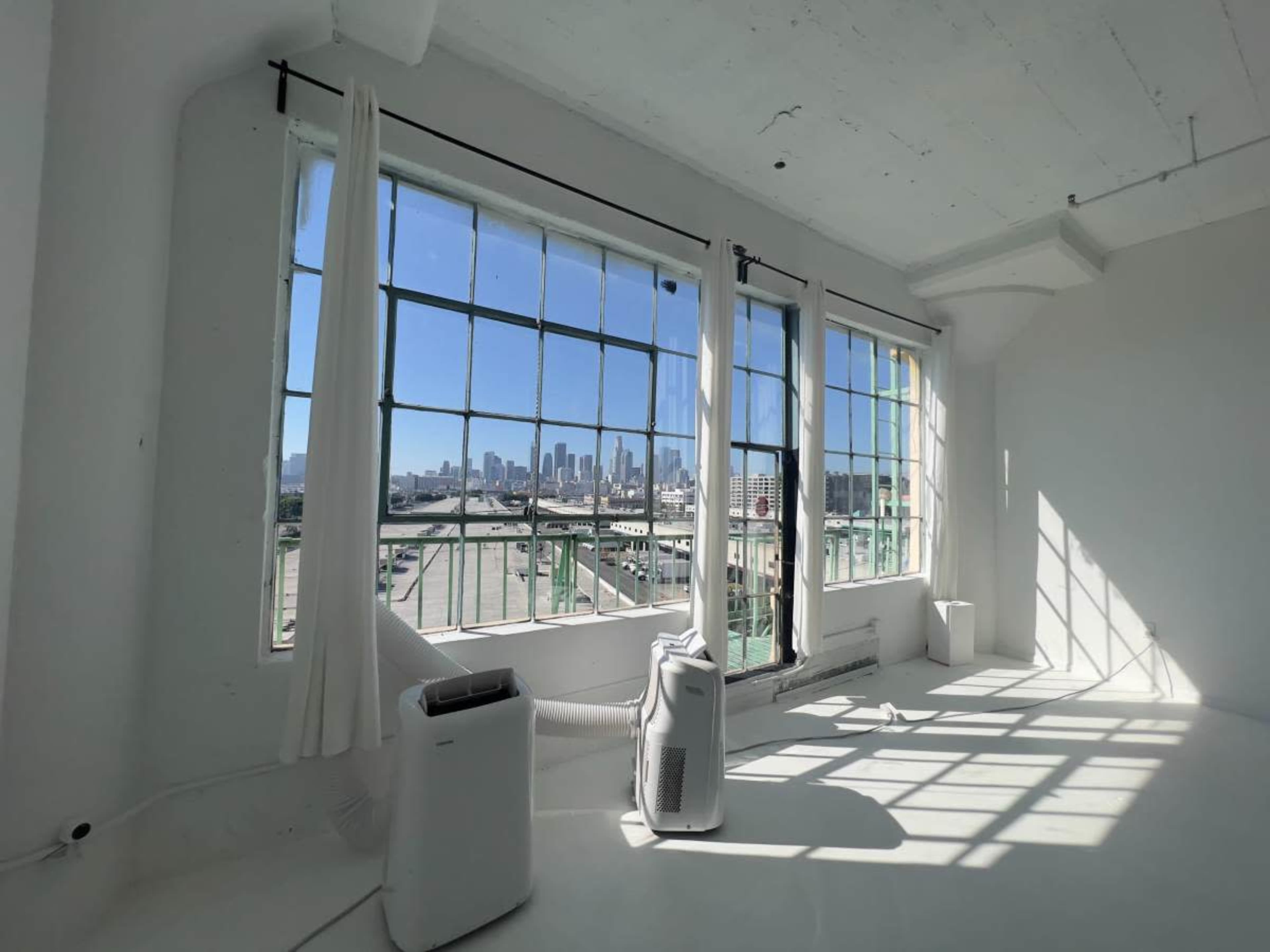 A bright, empty room with large windows overlooks a city skyline, featuring white walls and minimal furnishings.