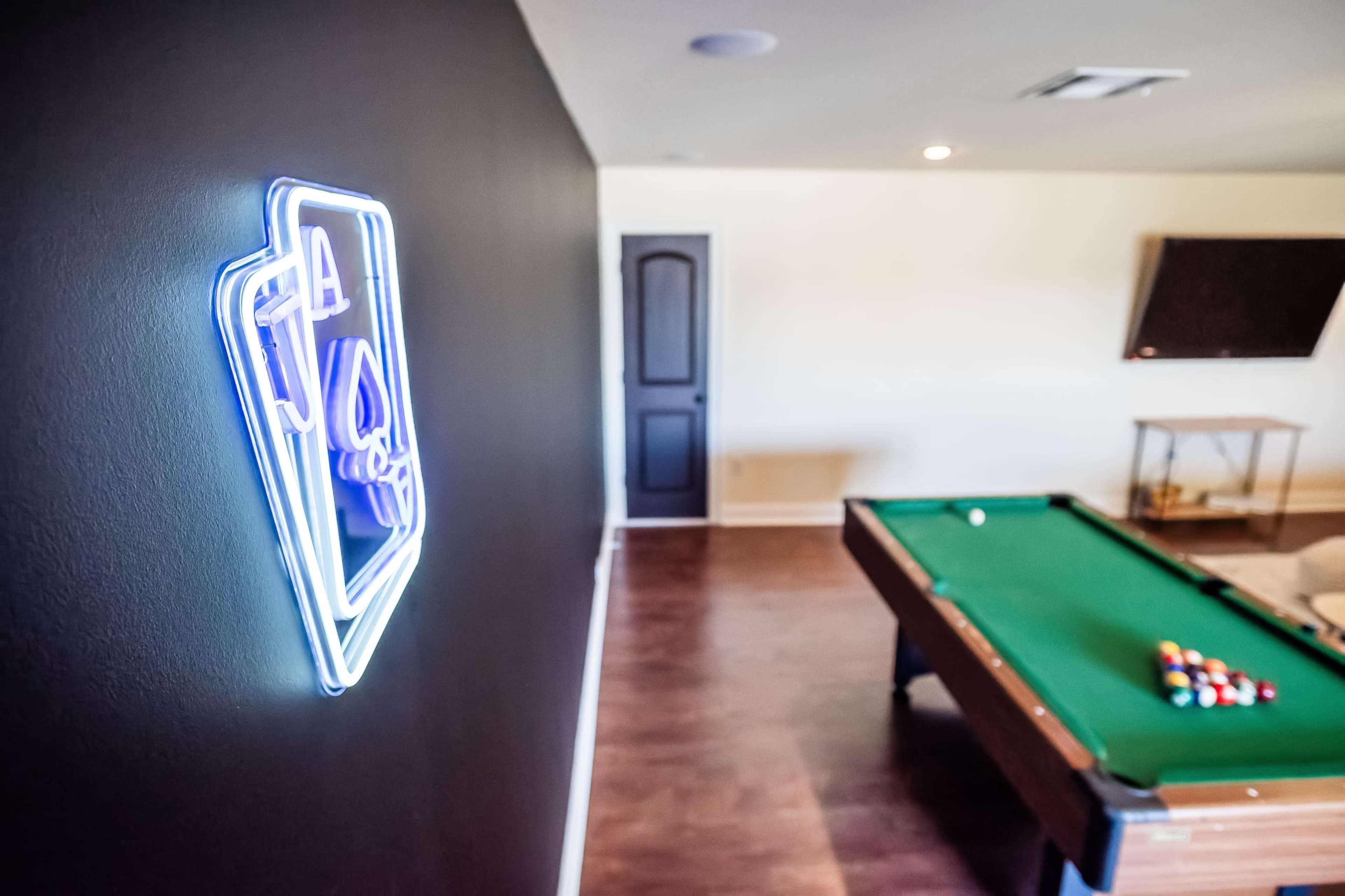 A game room with a billiard table, a neon sign on the wall, and a closed door in the background.