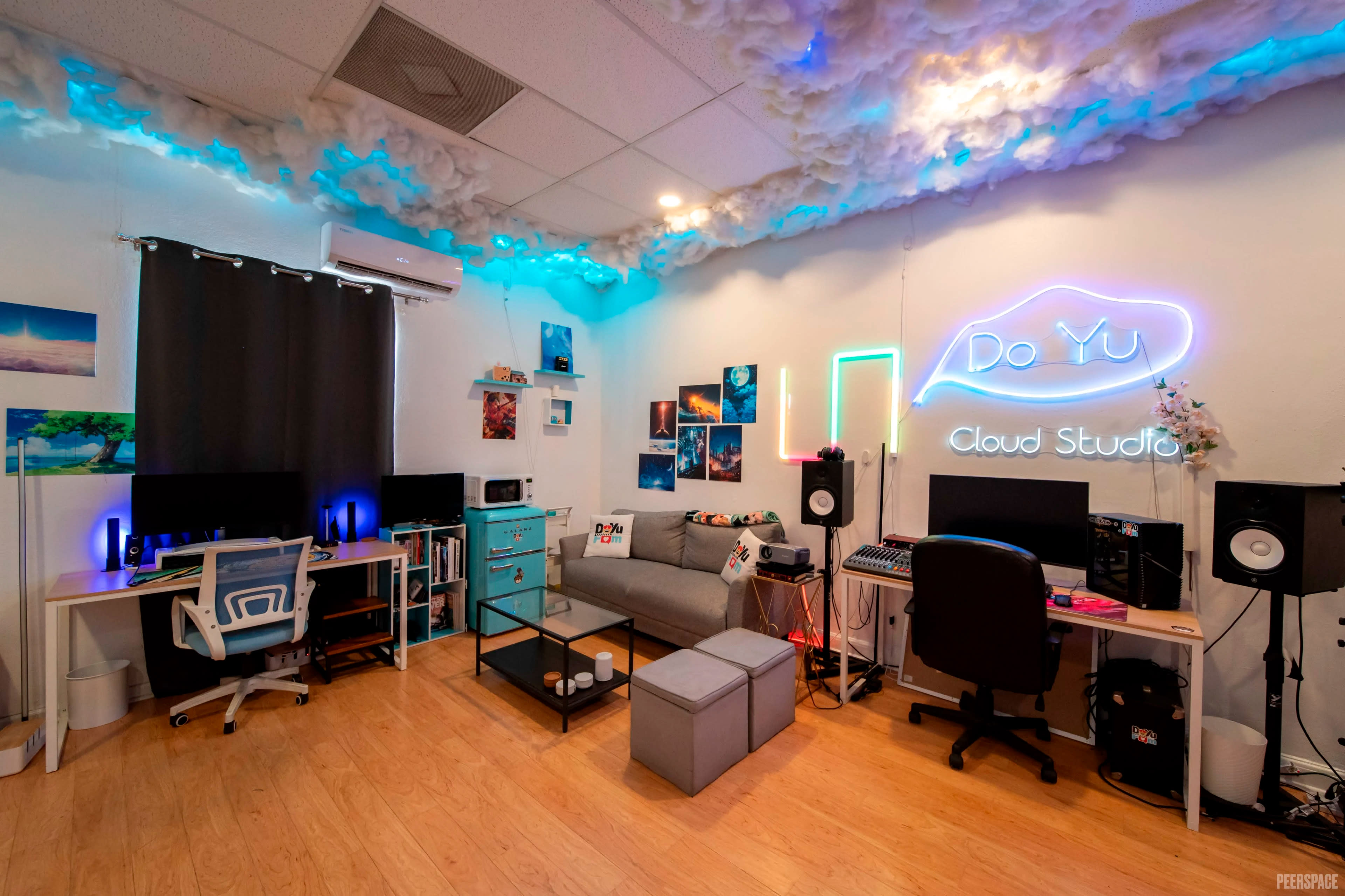 The image shows a creative studio space featuring two desks with computers, a couch, and colorful lighting, accented by cloud-like decorations on the ceiling.