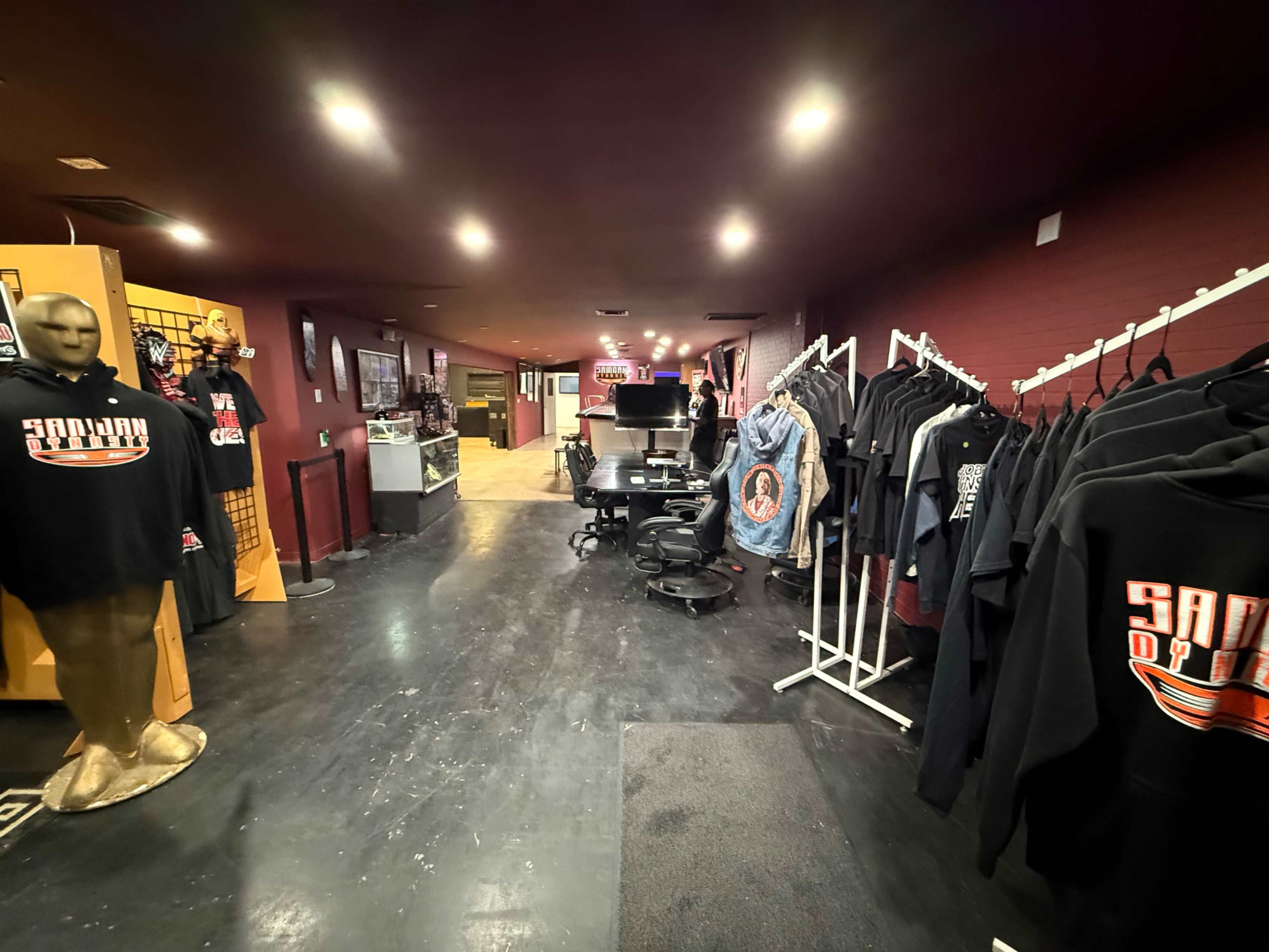 The image shows an interior view of a retail space with a row of clothing displays on one side and a workspace with chairs and equipment on the other.