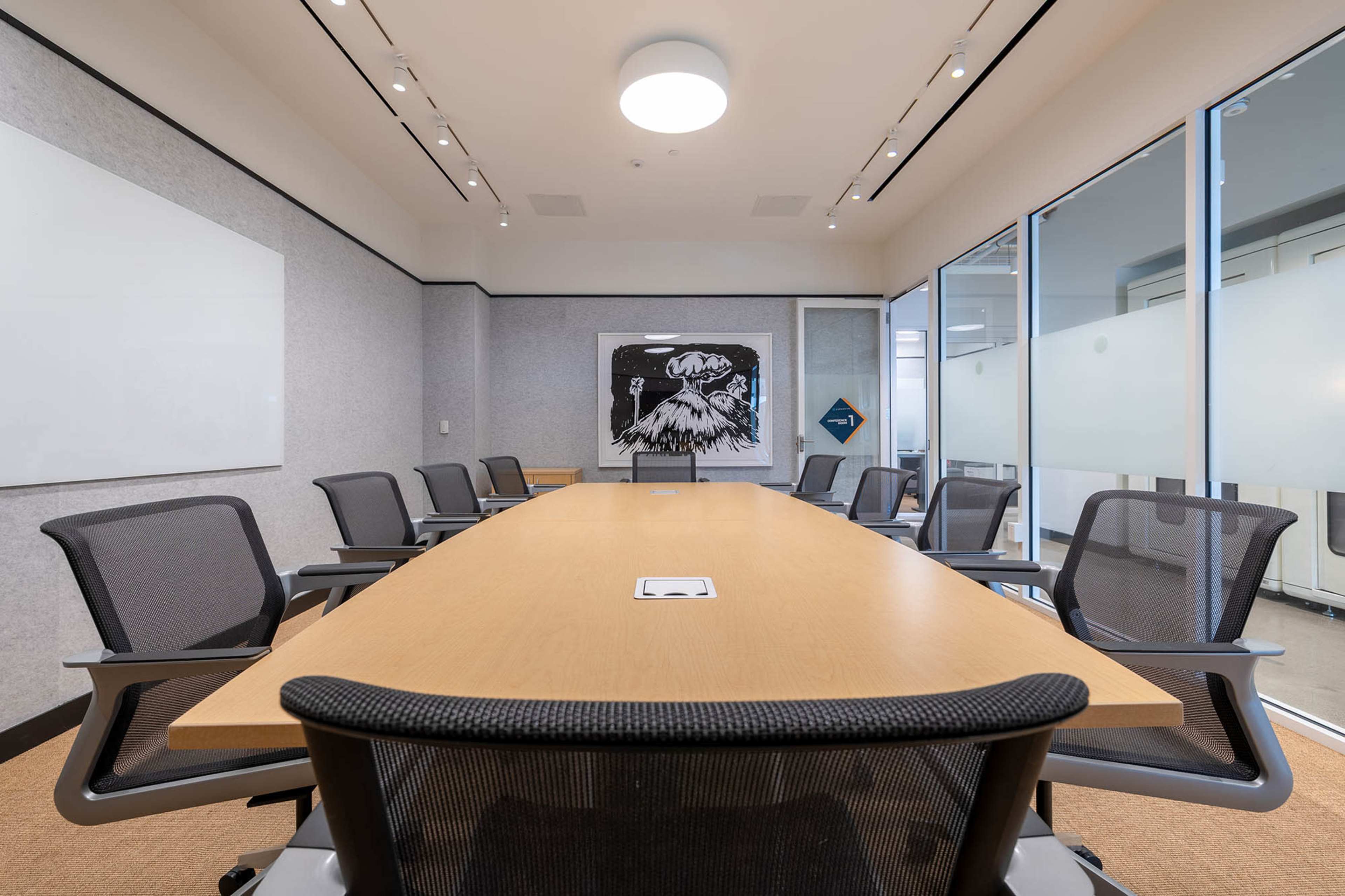 A spacious conference room features a long wooden table with ergonomic chairs, illuminated by ceiling lights, and a large artwork displayed on the wall.