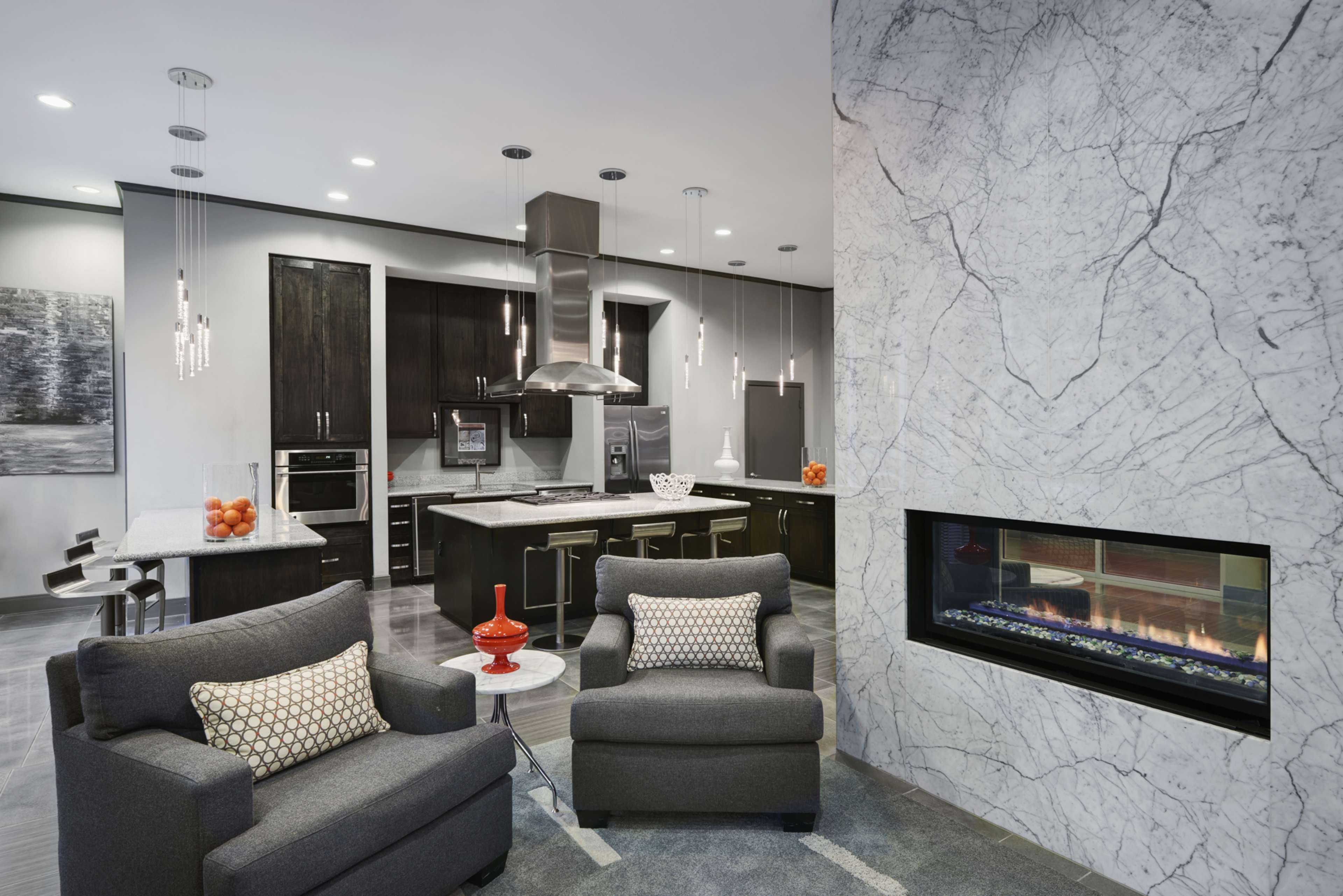 A modern kitchen and living area features gray armchairs, a marble fireplace, and sleek cabinetry with overhead lighting.