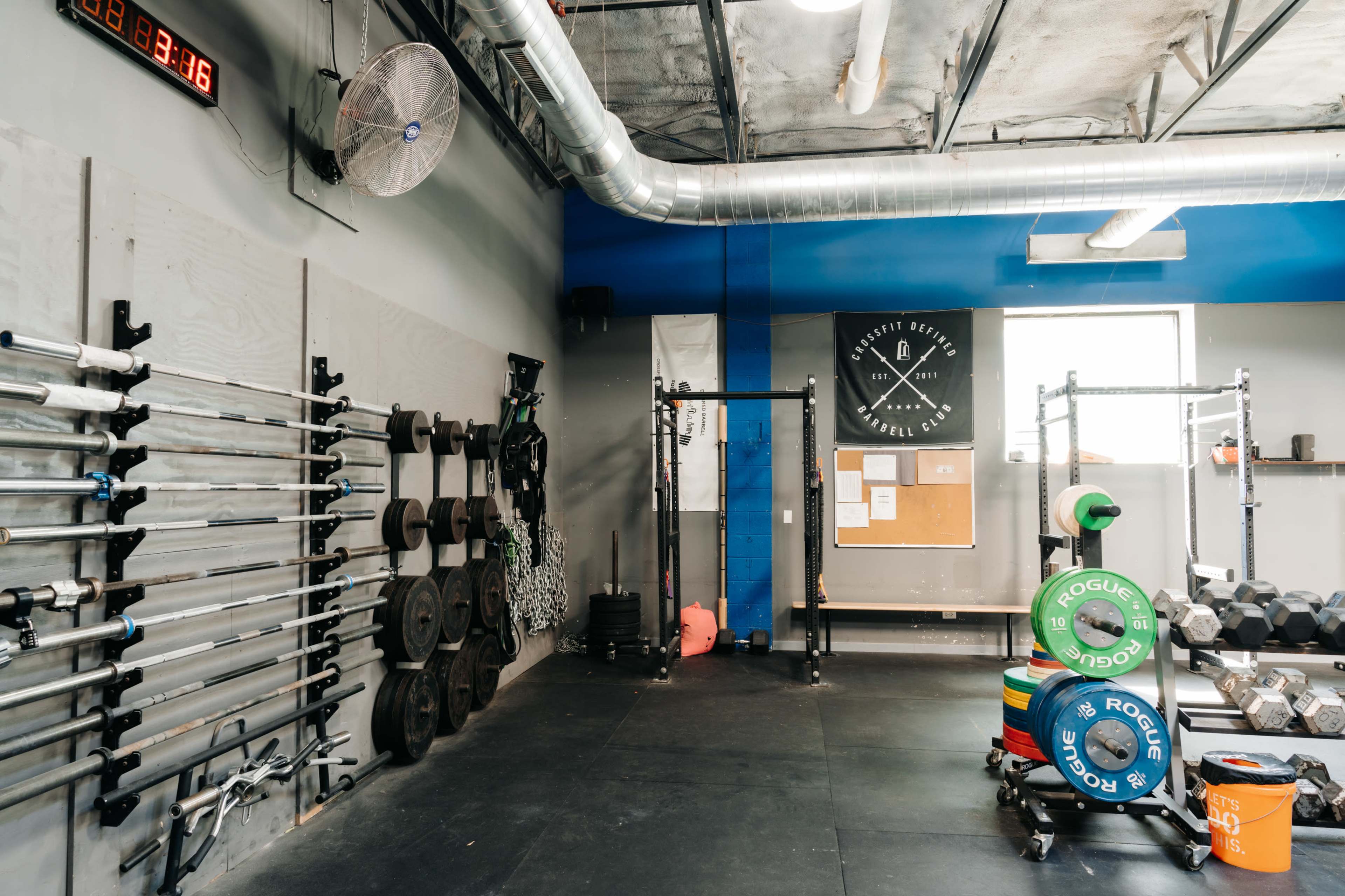 The image shows a gym interior with a wall-mounted clock, weightlifting bars, plates, a squat rack, and dumbbells arranged on a rubber floor.