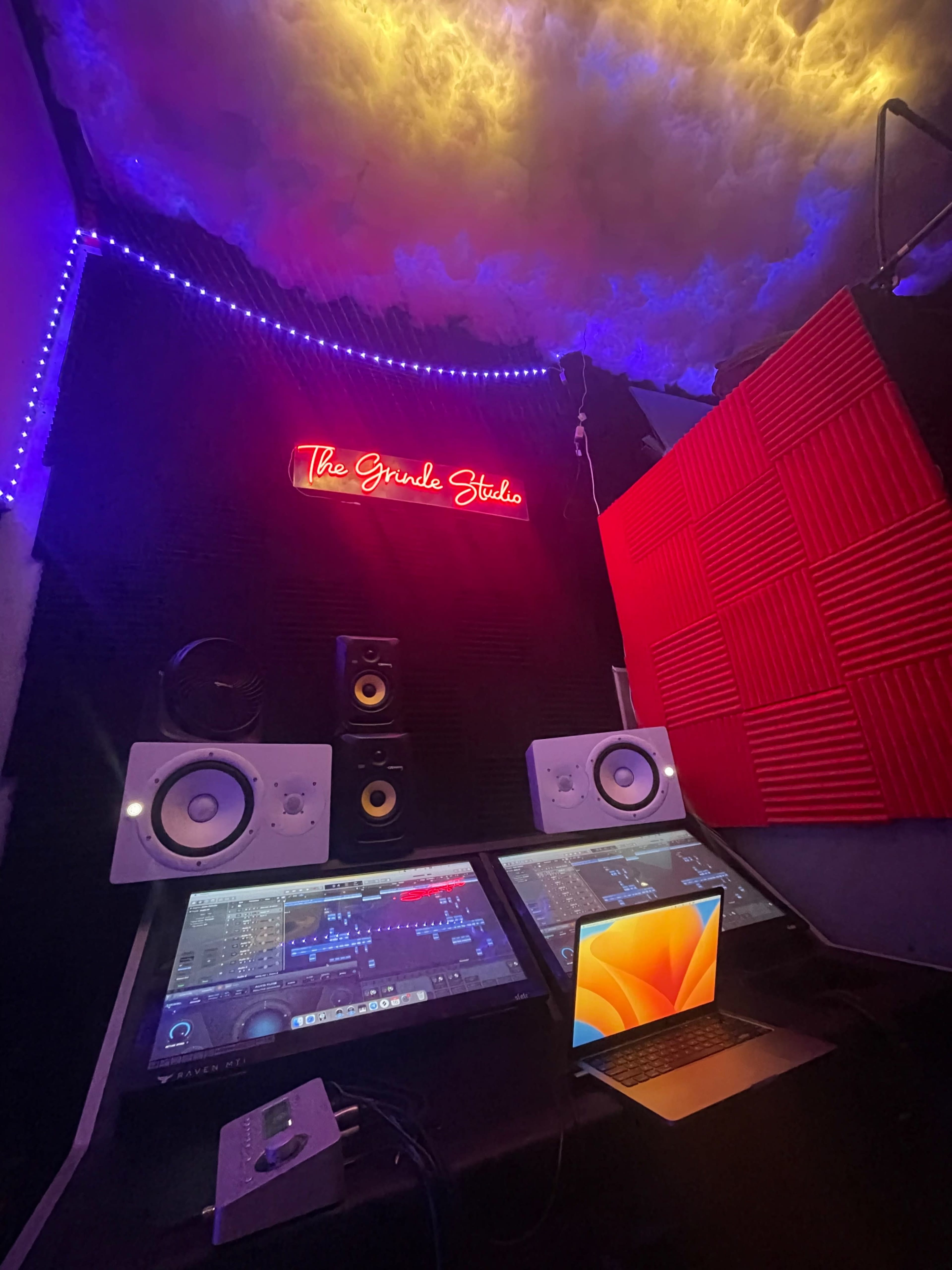 A music production studio featuring two monitors, speakers, and an illuminated sign that reads "The Grind Studio."