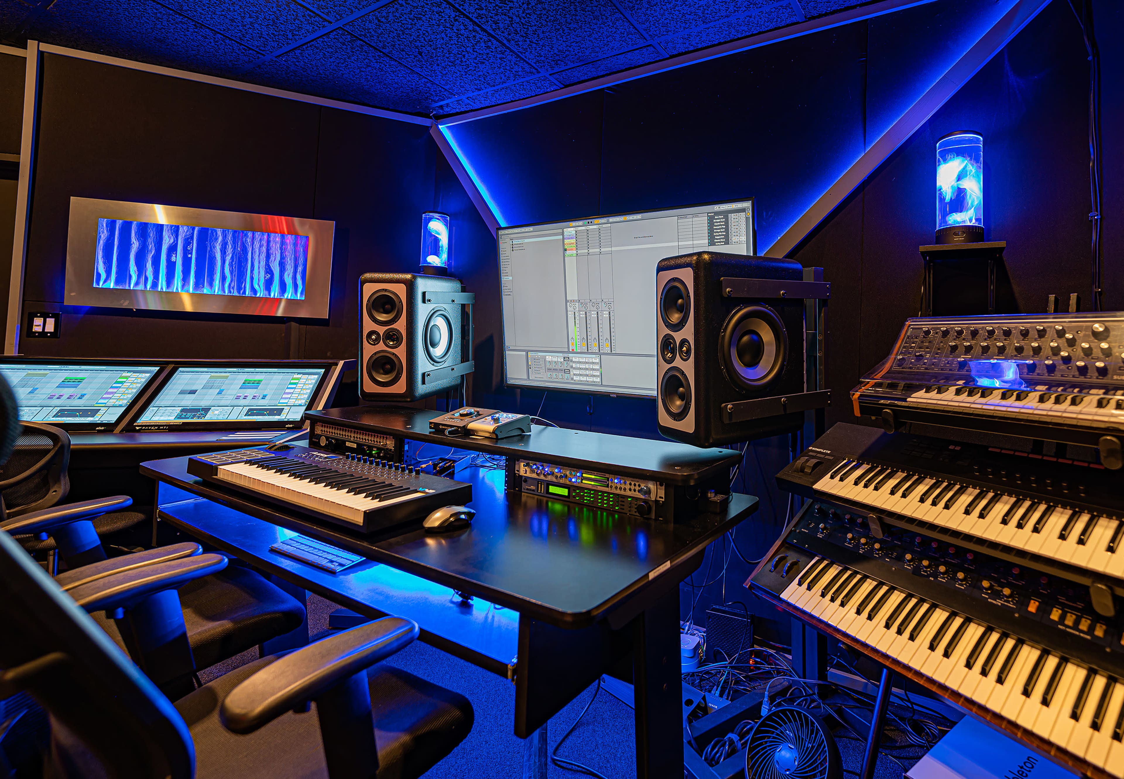 A modern music production studio equipped with a computer, mixing consoles, speakers, and multiple keyboards surrounded by blue lighting.