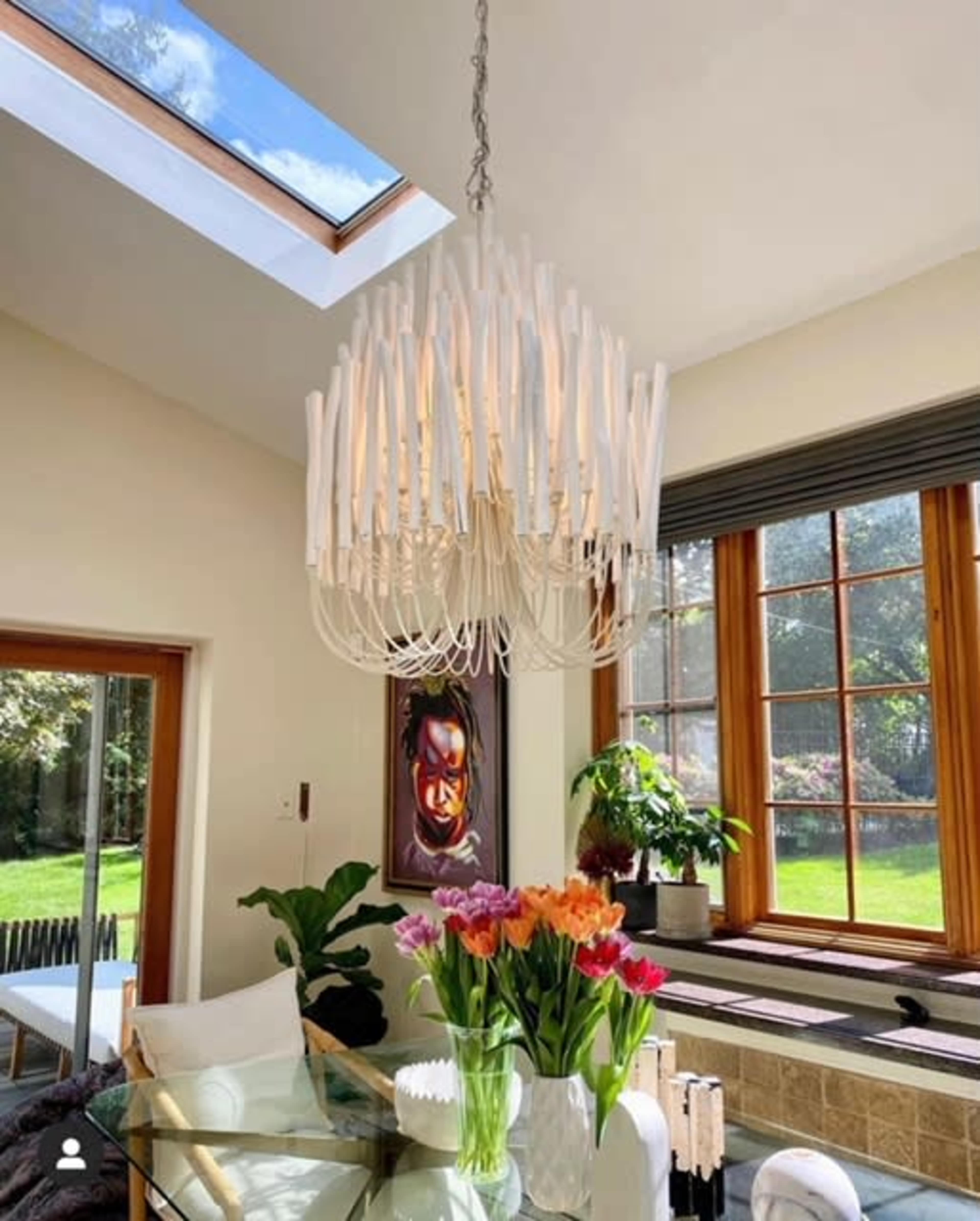 A modern chandelier hangs above a glass dining table adorned with a bouquet of colorful flowers, while large windows illuminate the room with natural light.