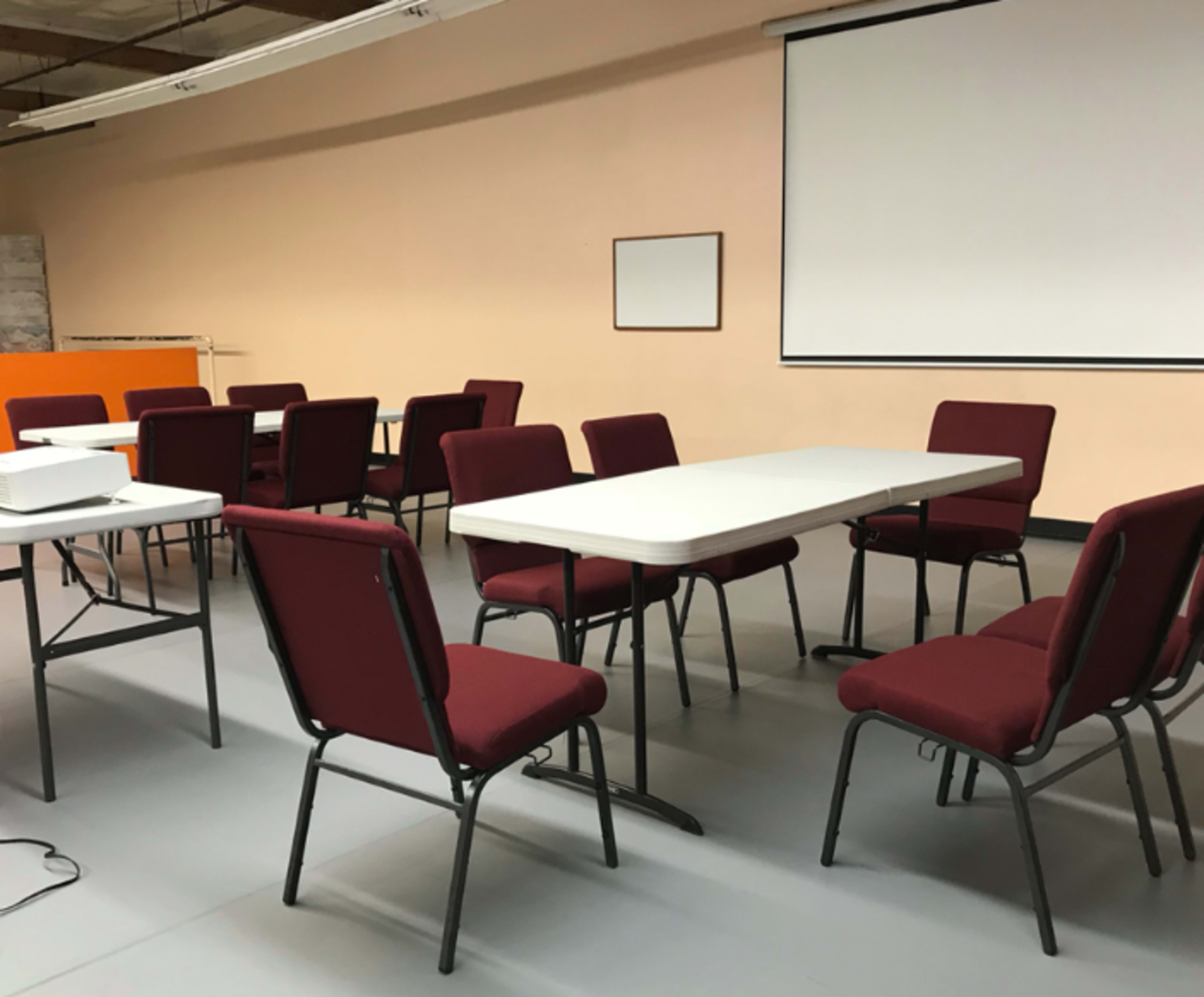 A meeting room is arranged with several chairs and tables, featuring a projector and a blank screen.
