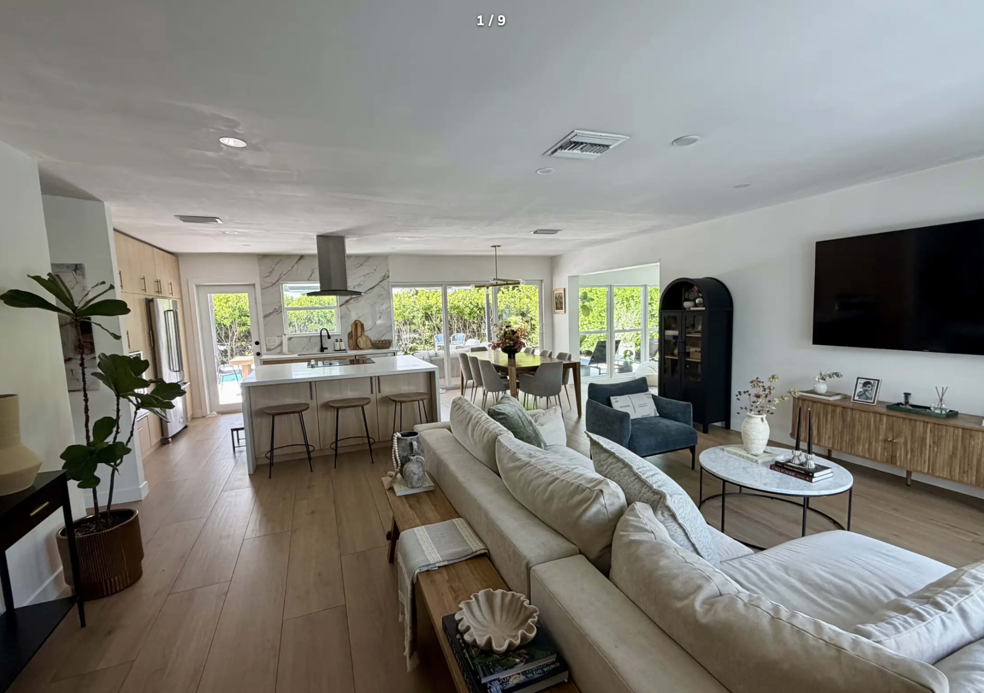 The image shows a spacious, well-lit living area with a sectional sofa, dining table, and a modern kitchen in an open-concept layout.