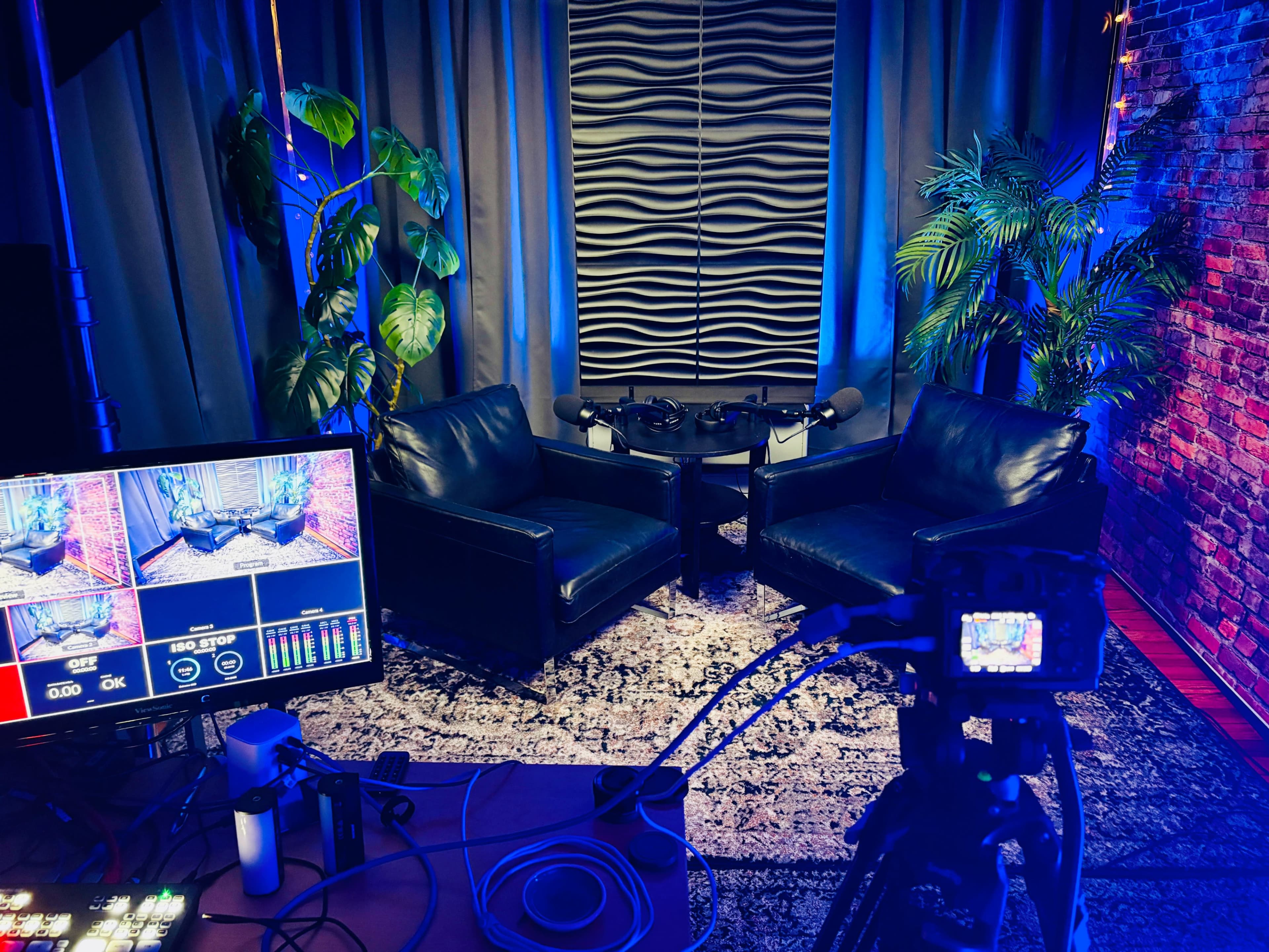 The image shows a recording studio setup with two black chairs facing each other, surrounded by decorative plants and a desk equipped with recording equipment and a monitor.