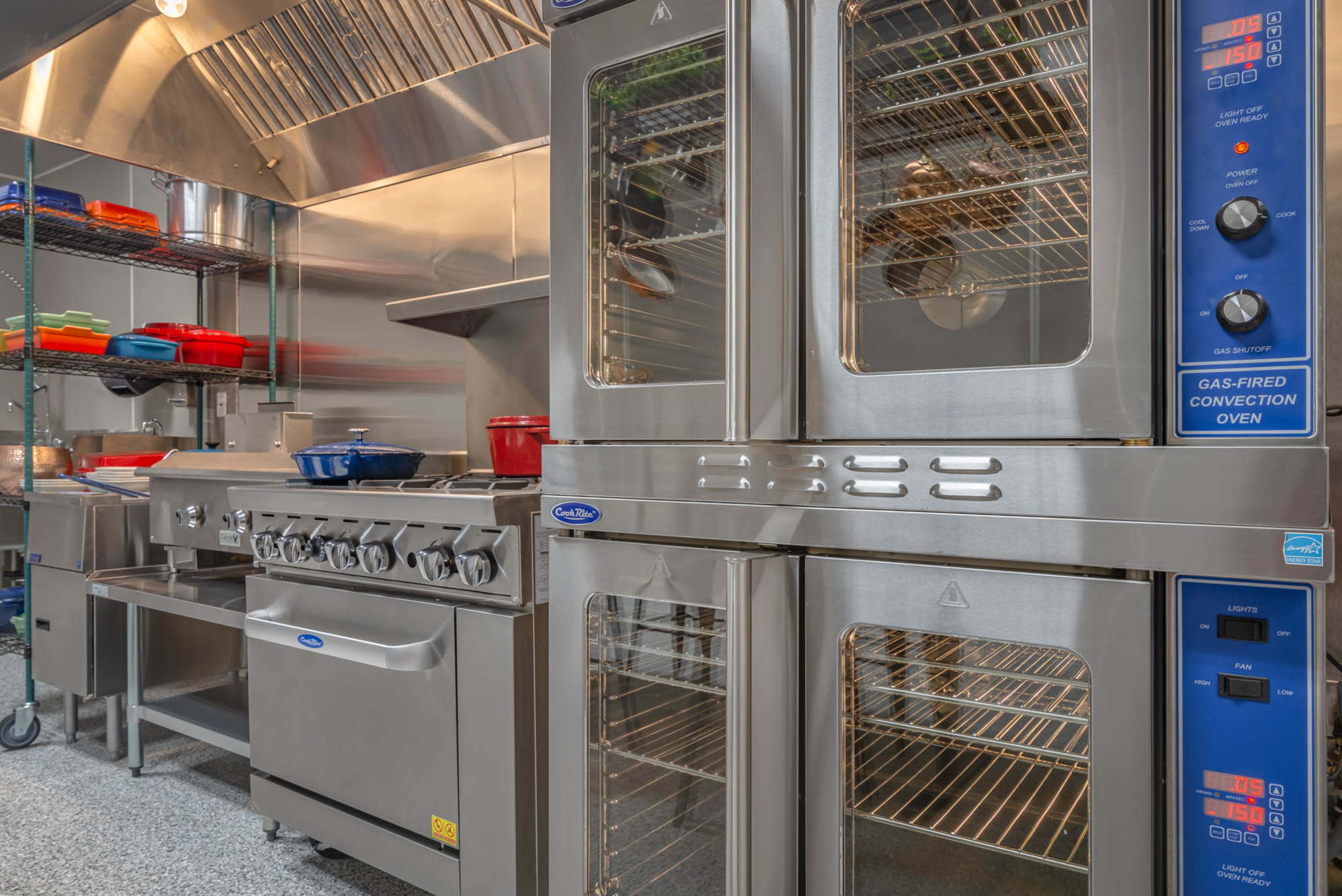 The image shows a modern commercial kitchen equipped with stainless steel ovens, a gas range, and organized shelving.