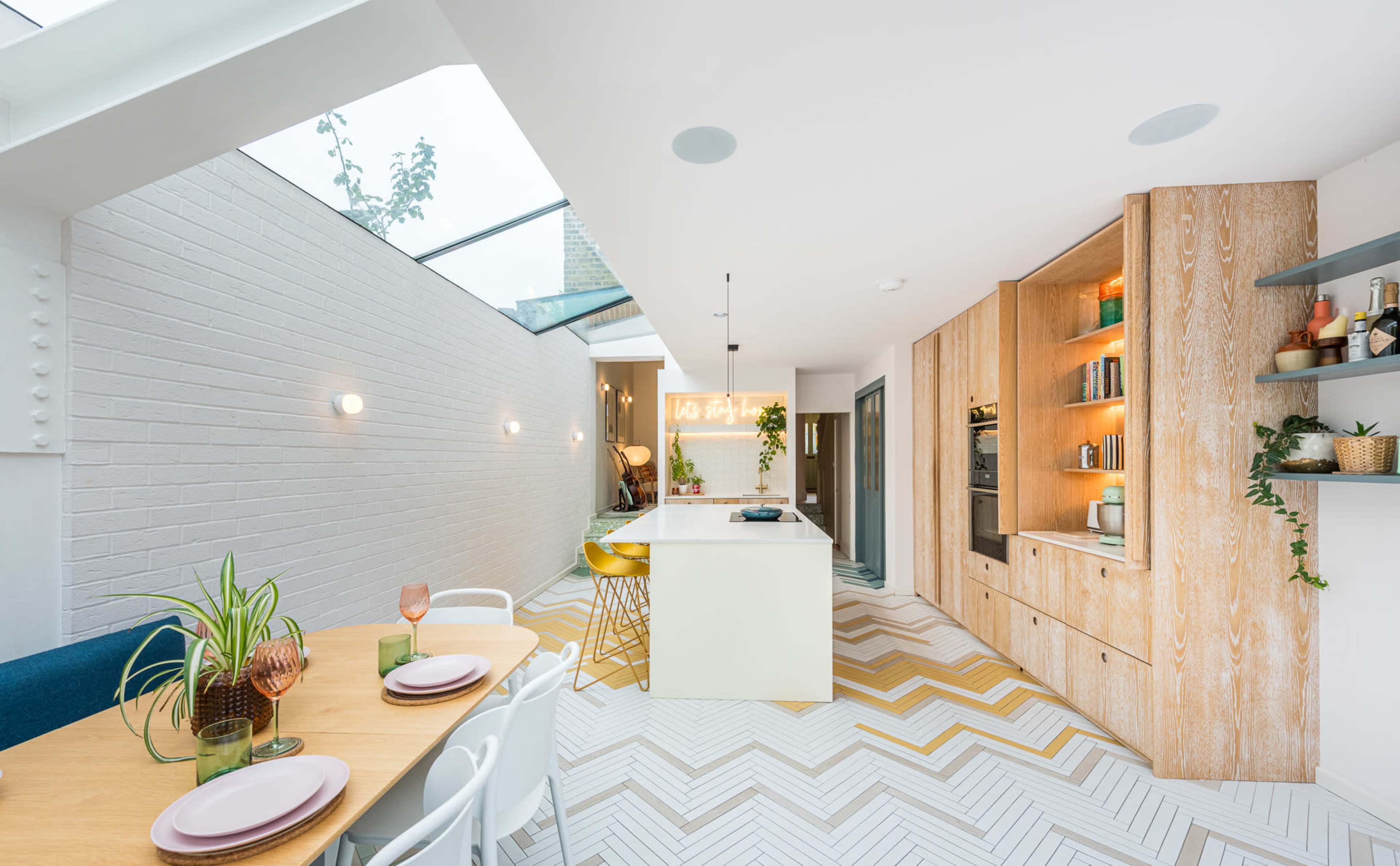 A modern, open-concept kitchen and dining area features a long dining table, colorful chairs, and a center island with a skylight above, showcasing geometric patterned flooring.