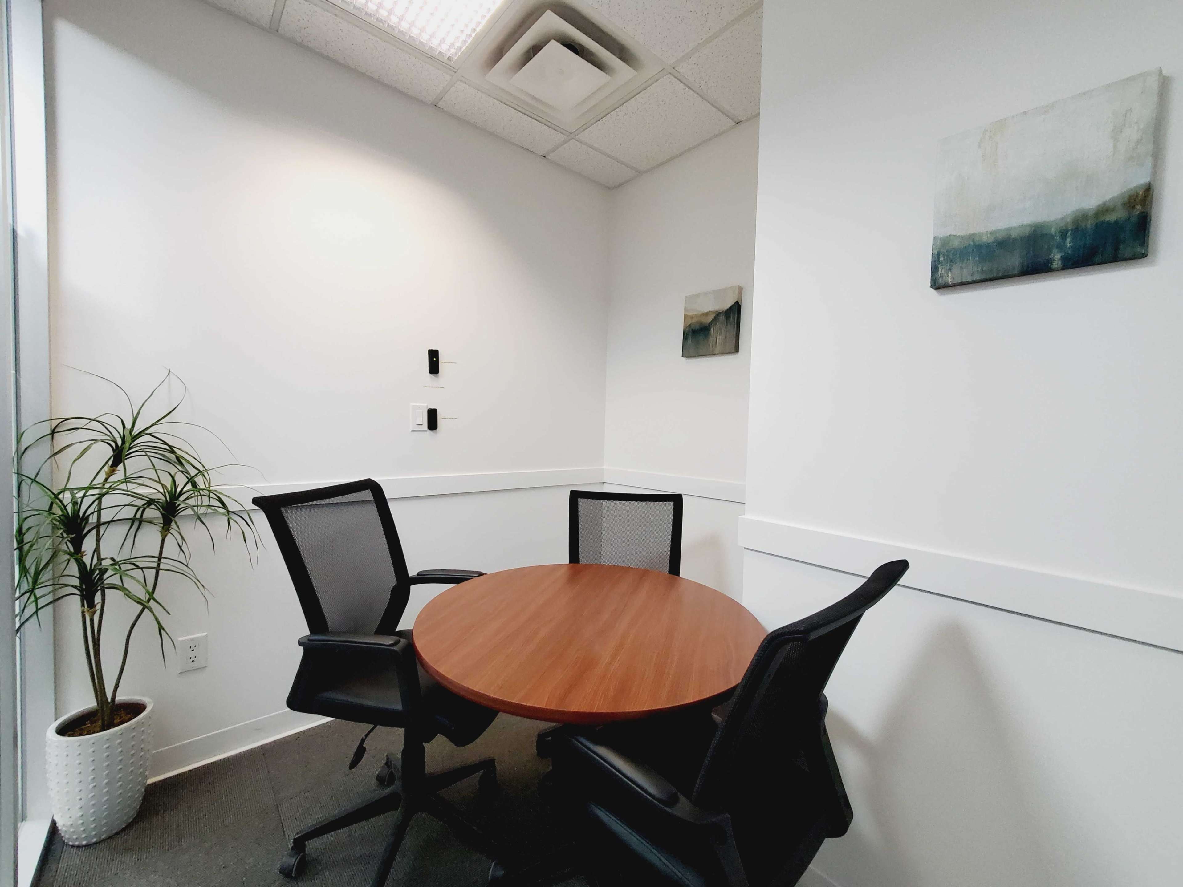 A small meeting room features a round wooden table surrounded by two black chairs, with a plant in a pot and framed artwork on the walls.