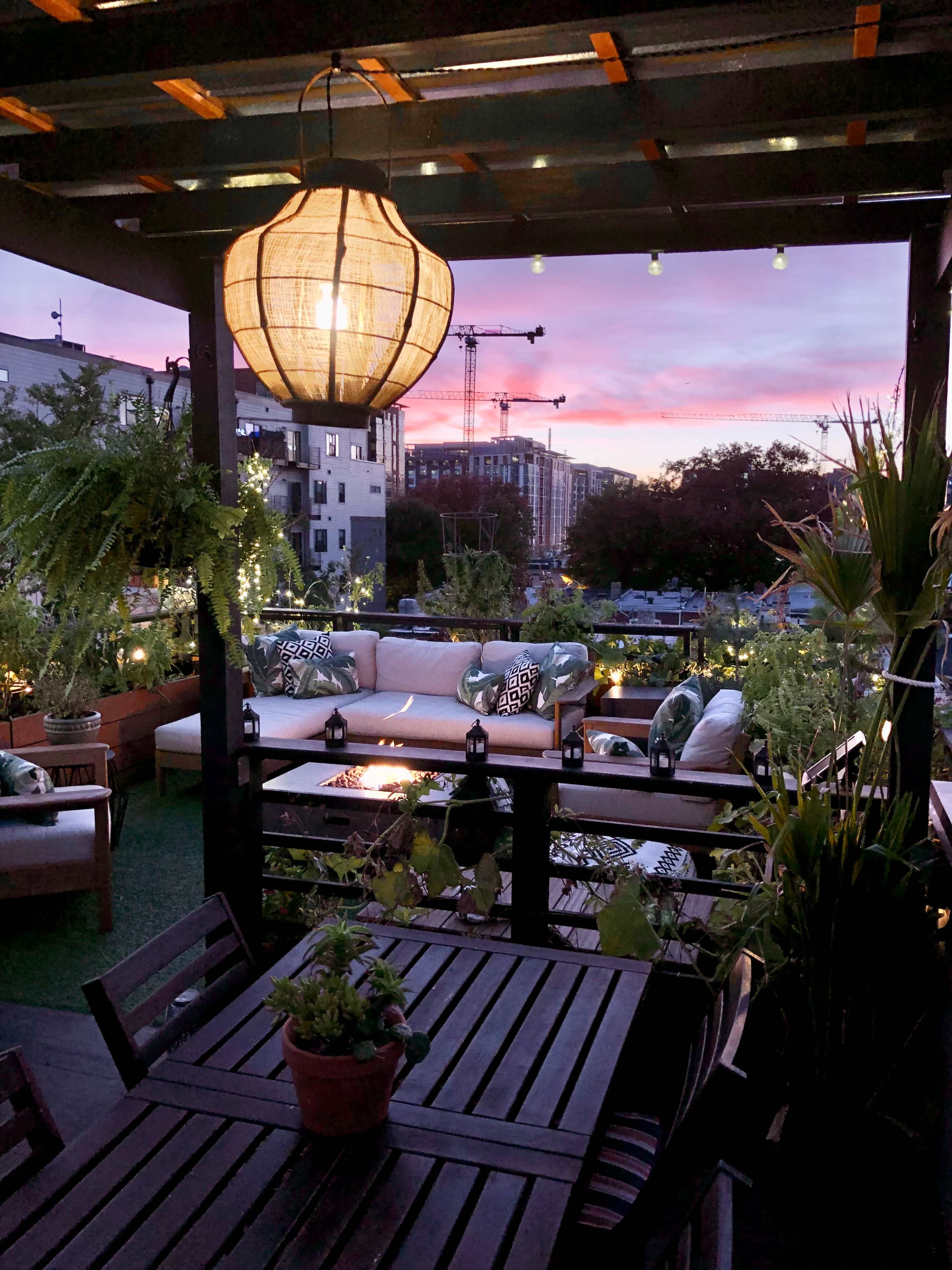 A rooftop terrace features a lantern, comfortable seating, and potted plants under a colorful sunset sky.