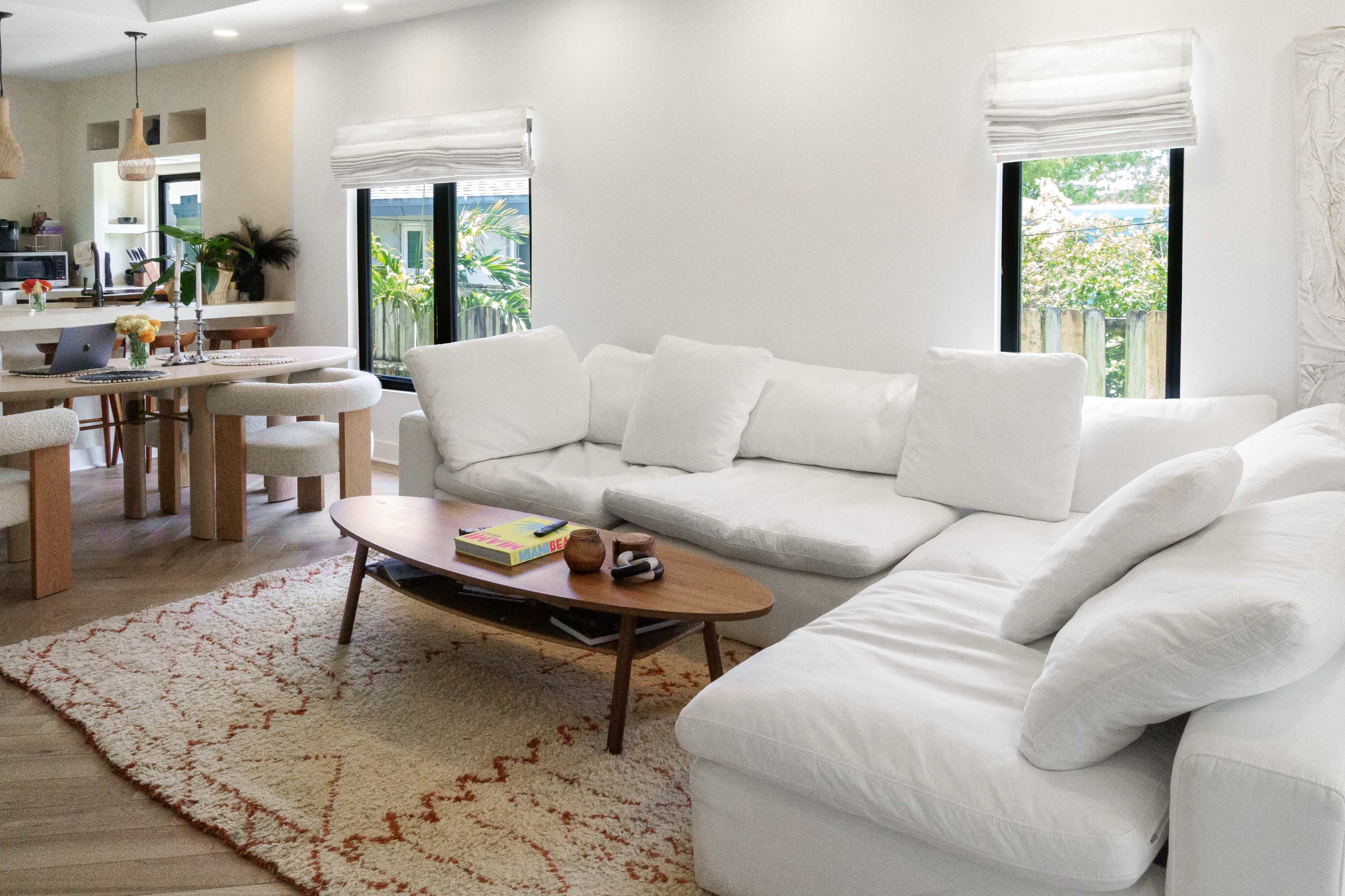 A bright living room features a white sectional sofa, a wooden coffee table, and a dining area with a round table near large windows surrounded by greenery.