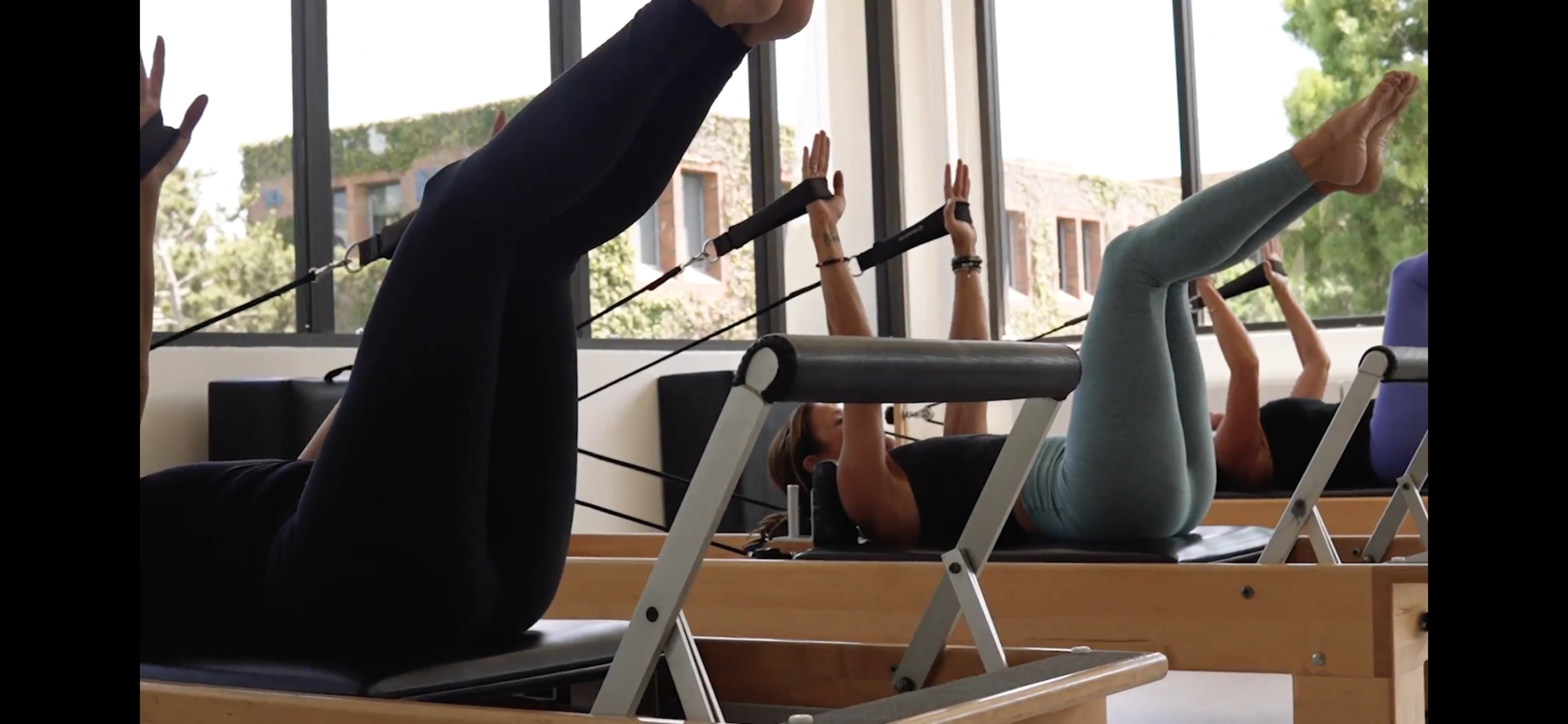Two individuals perform Pilates exercises on reformer machines in a well-lit studio with large windows.