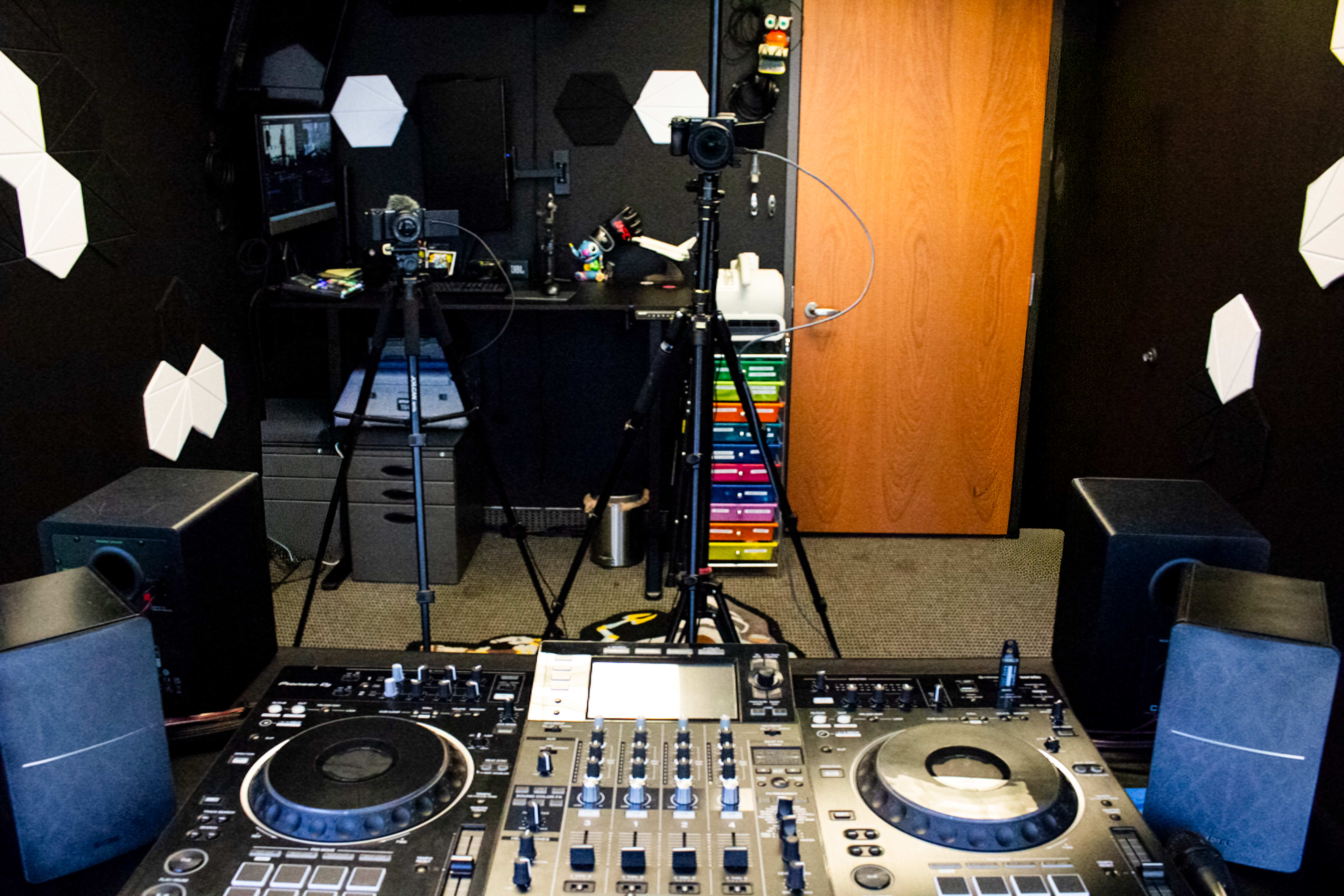 The image shows a DJ setup with turntables and speakers in front of a wall featuring geometric shapes, with cameras and equipment visible in the background.