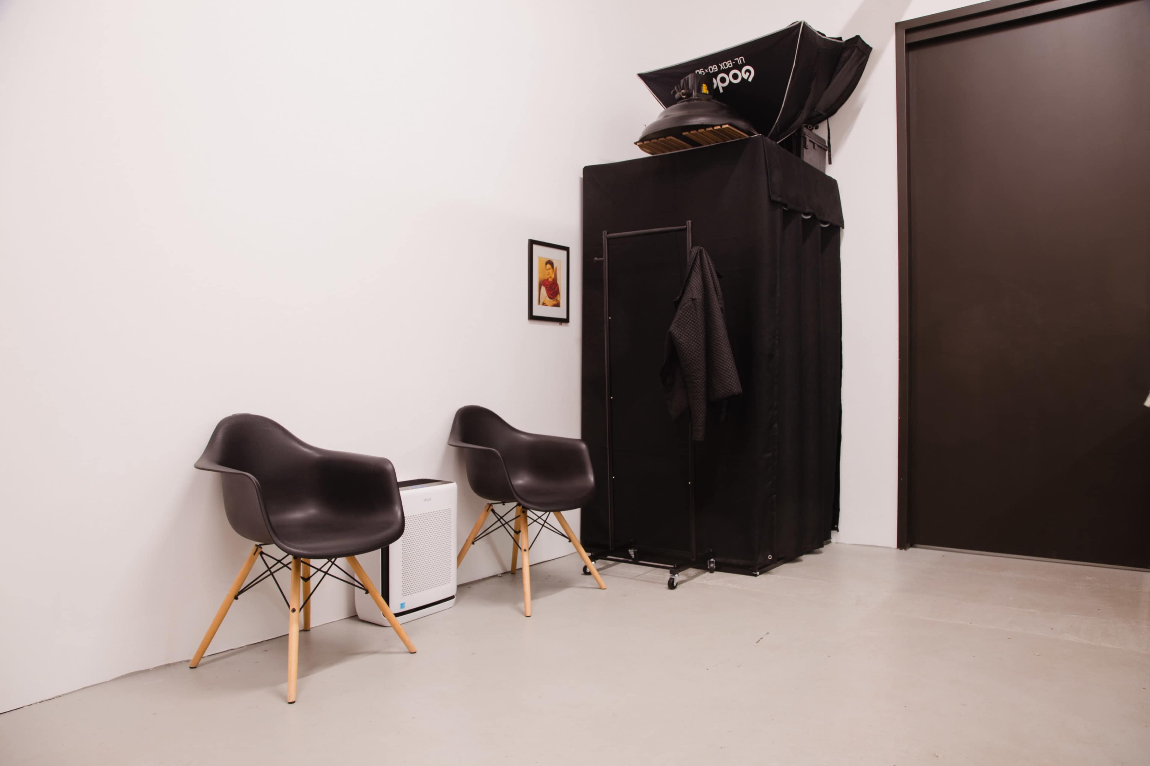 The image depicts a minimalistic room with two black chairs, a small white refrigerator, and a portable photography backdrop on one side.