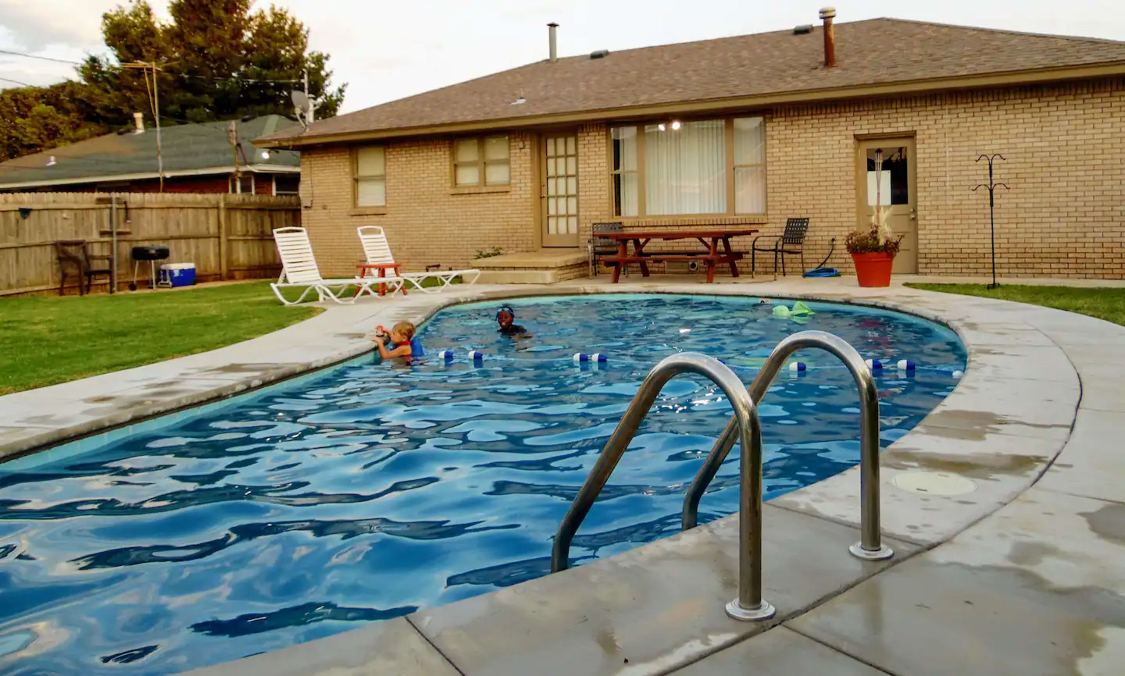 A backyard swimming pool with two children playing in the water beside a brick house and lawn furniture.