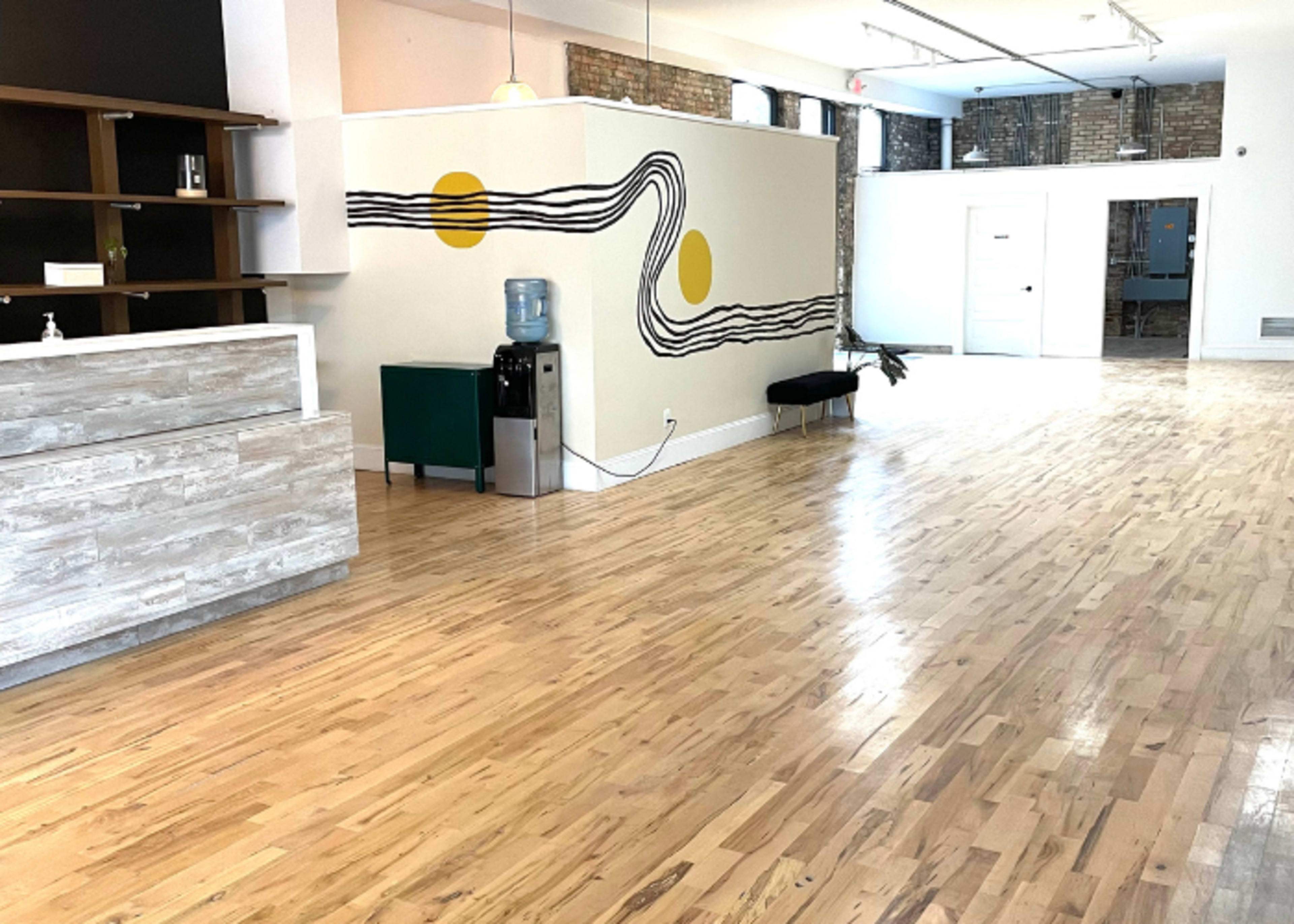 A spacious interior with light wooden flooring, a reception area featuring a counter, a green water cooler, and a wall decorated with abstract patterns and yellow circles.