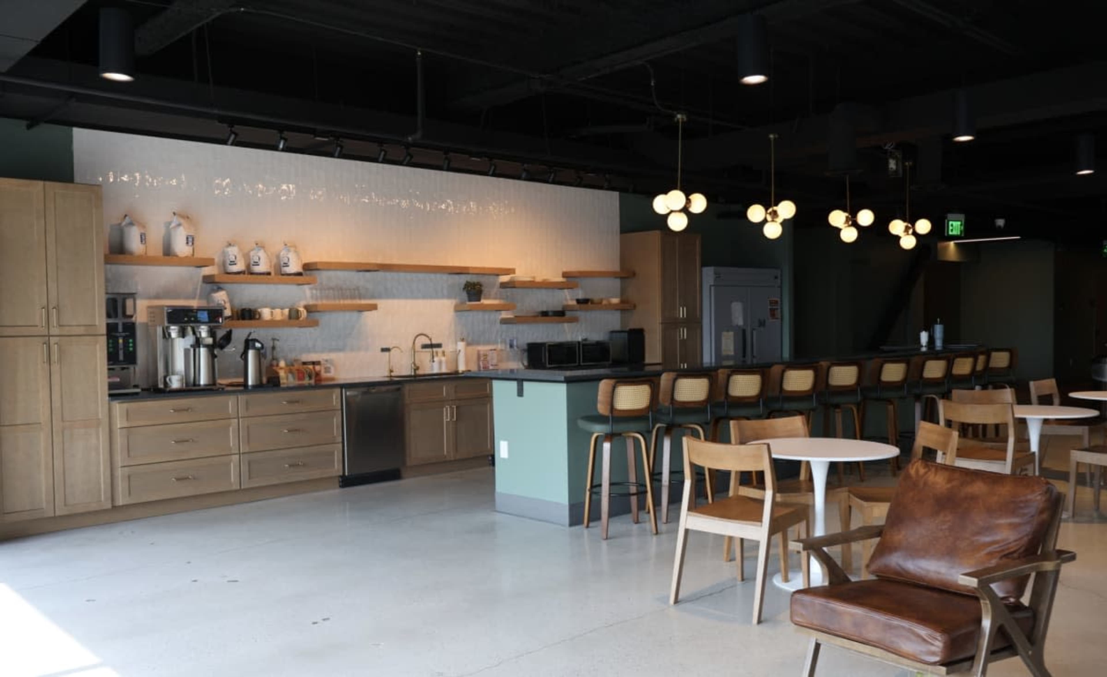 The image shows a modern coffee bar with wooden cabinetry, a countertop with various coffee machines, and seating including wooden chairs and tables in a spacious interior.