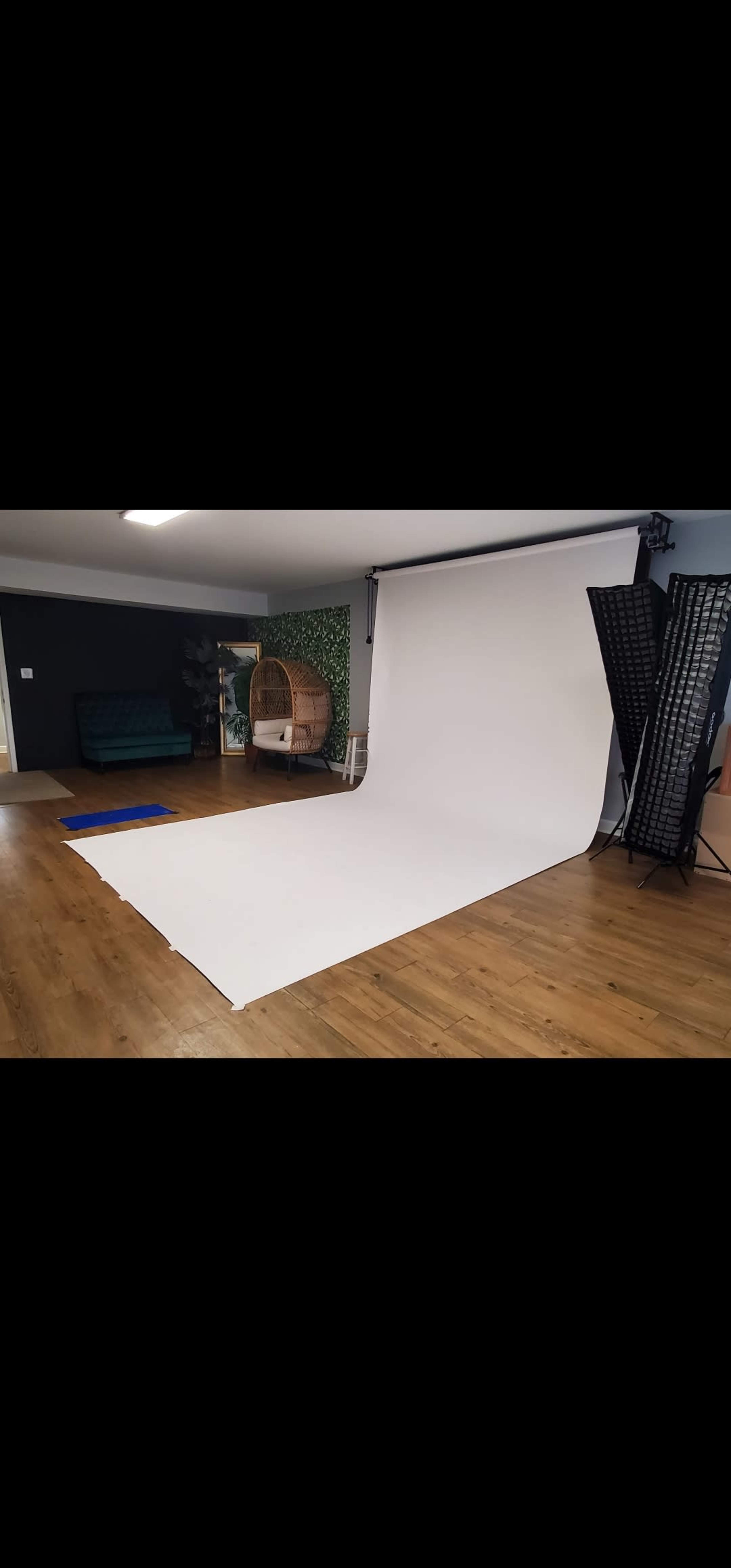 A photography studio with a white backdrop, wooden floor, a green couch, a decorative plant wall, and lighting equipment on the side.