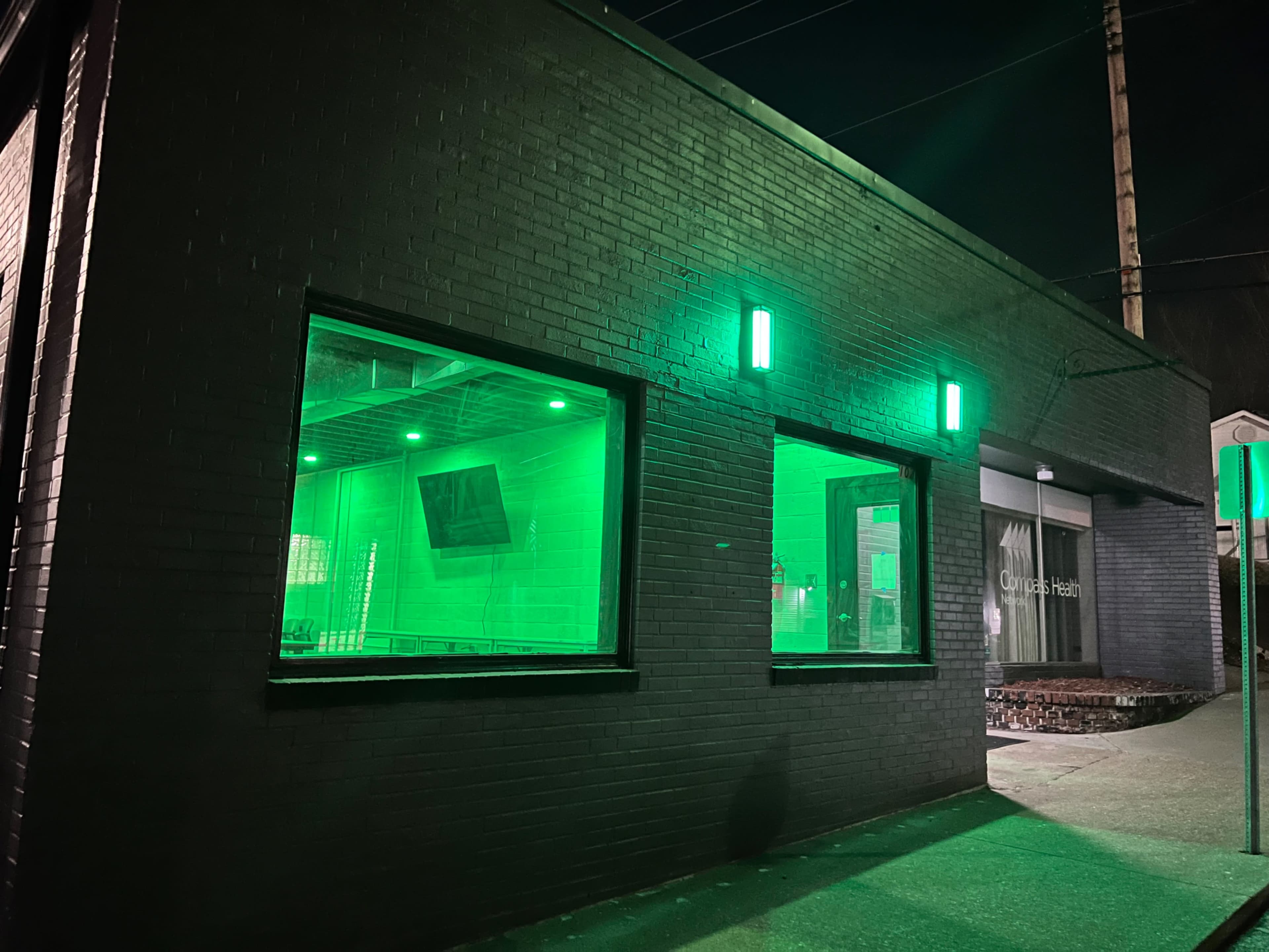 A dark building illuminated by green lights at night with large windows showing an interior space.