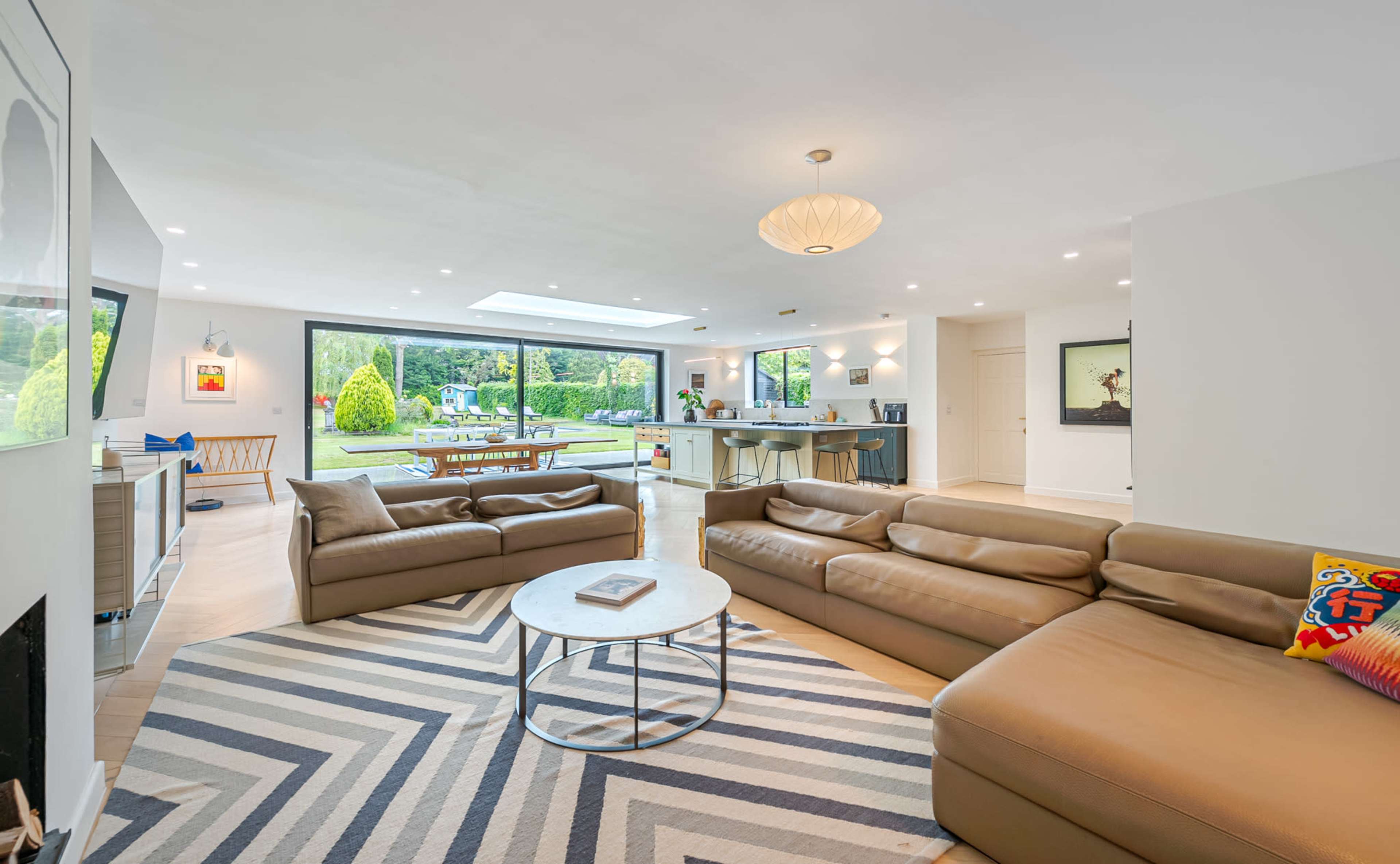 The image depicts a modern living room featuring two brown leather sofas, a circular coffee table, and large windows overlooking a backyard with greenery.