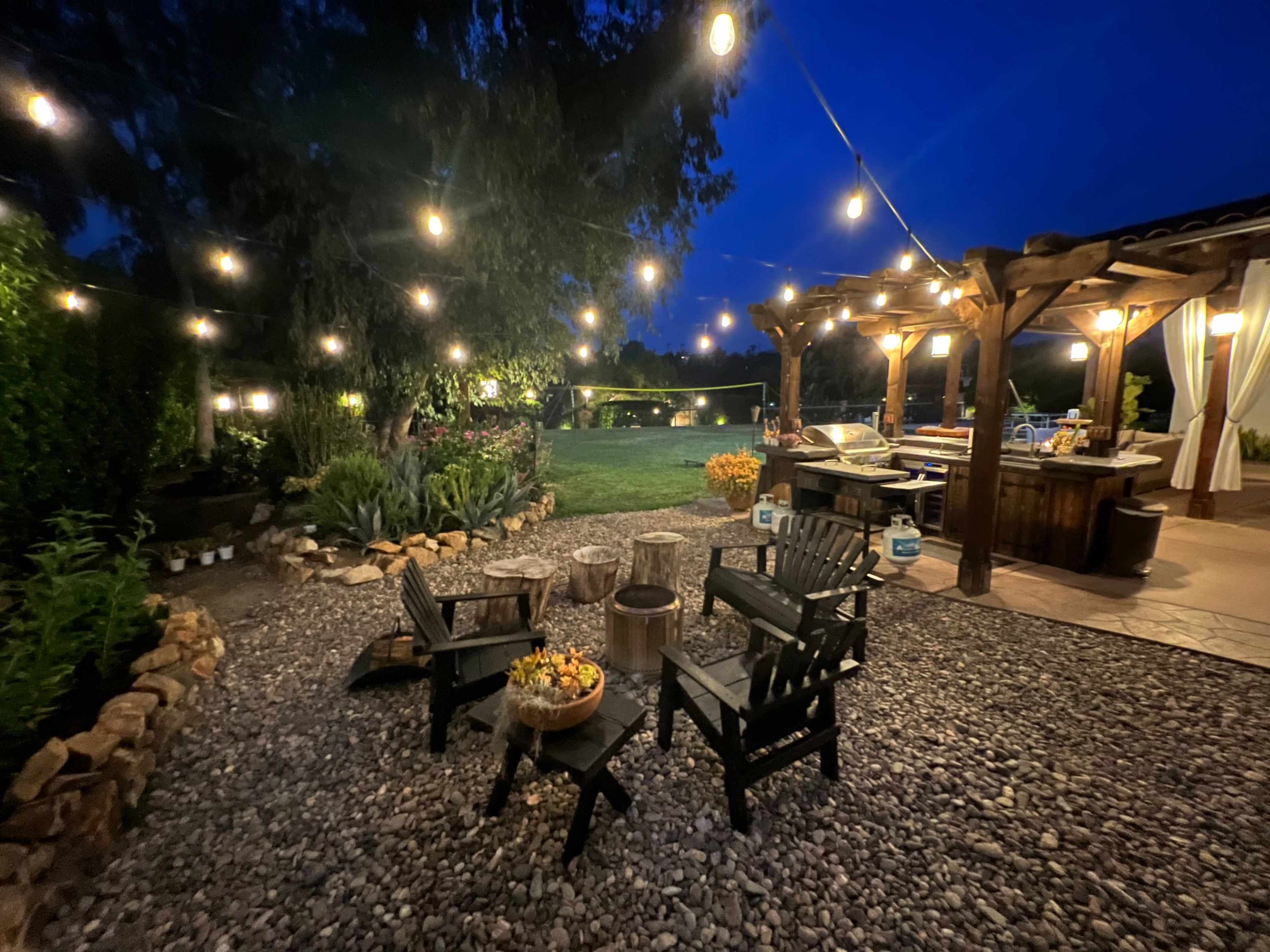 A cozy outdoor seating area with string lights overhead, featuring wooden chairs around a firepit and a rustic bar in the background.