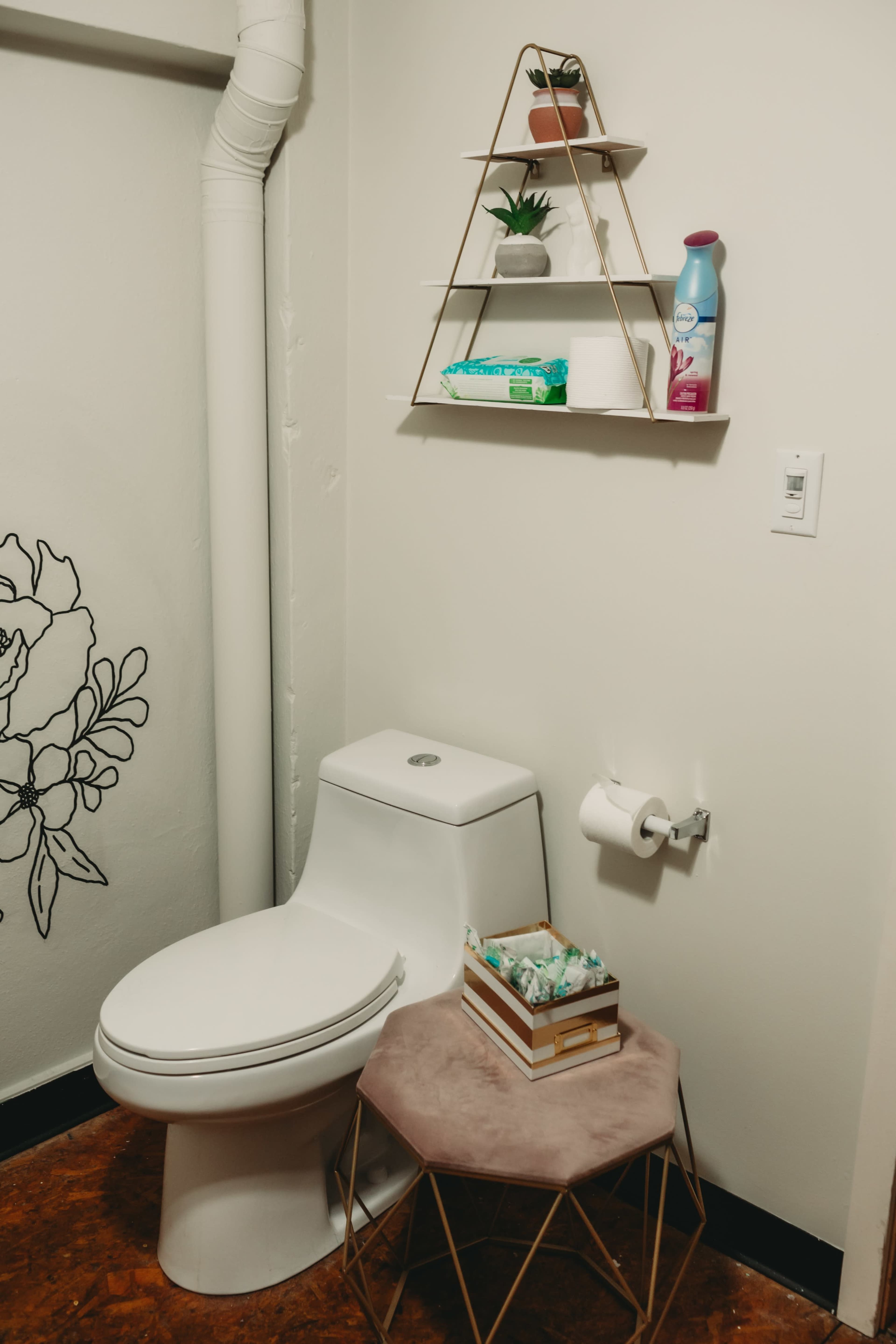 A bathroom corner with a white toilet, a small circular table holding toiletries, and a wall-mounted shelf displaying plants and cleaning supplies.