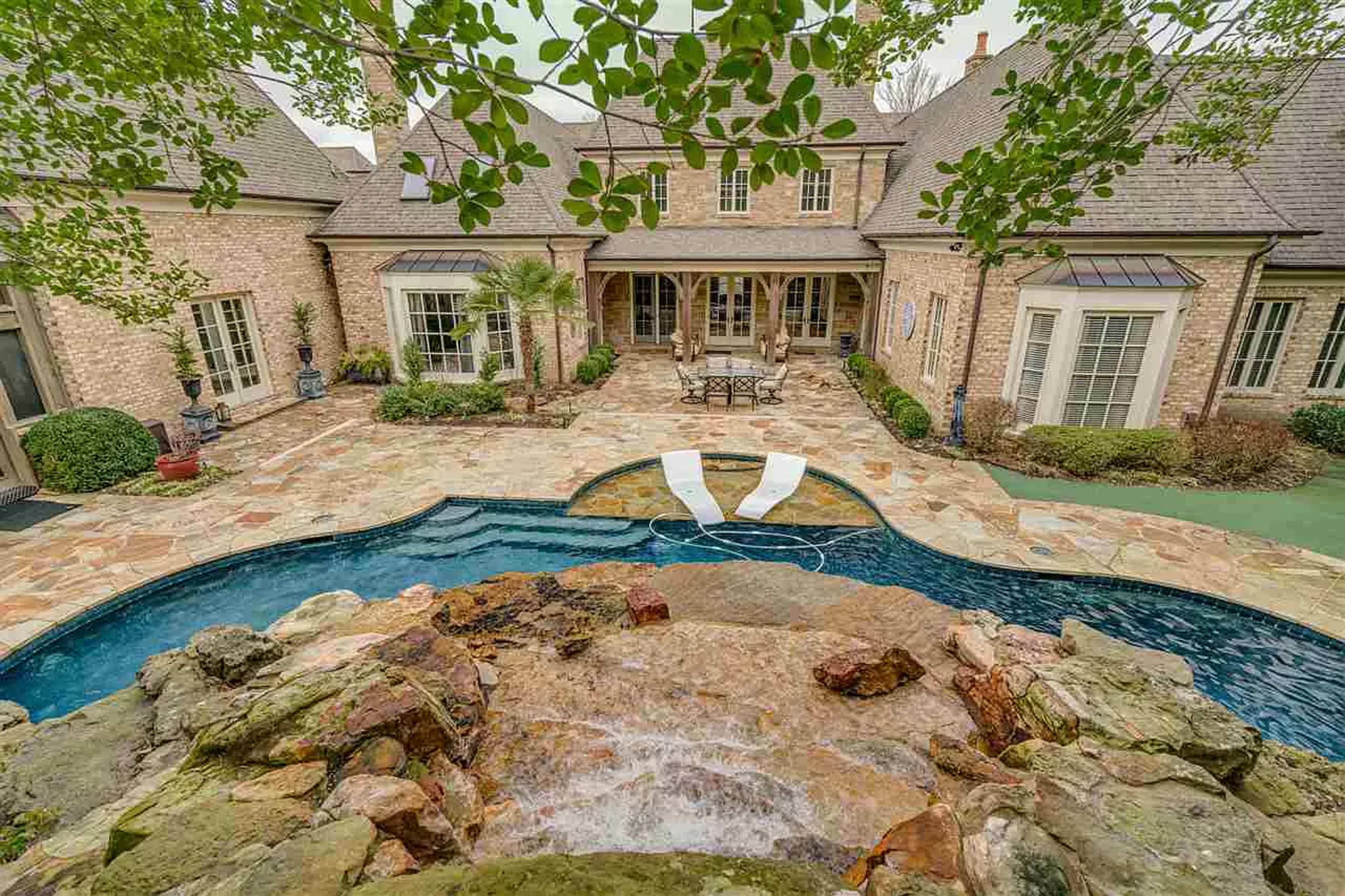 A stone patio surrounds a swimming pool with a waterfall feature and lounge chairs, framed by the exterior of a large brick house and landscaped greenery.