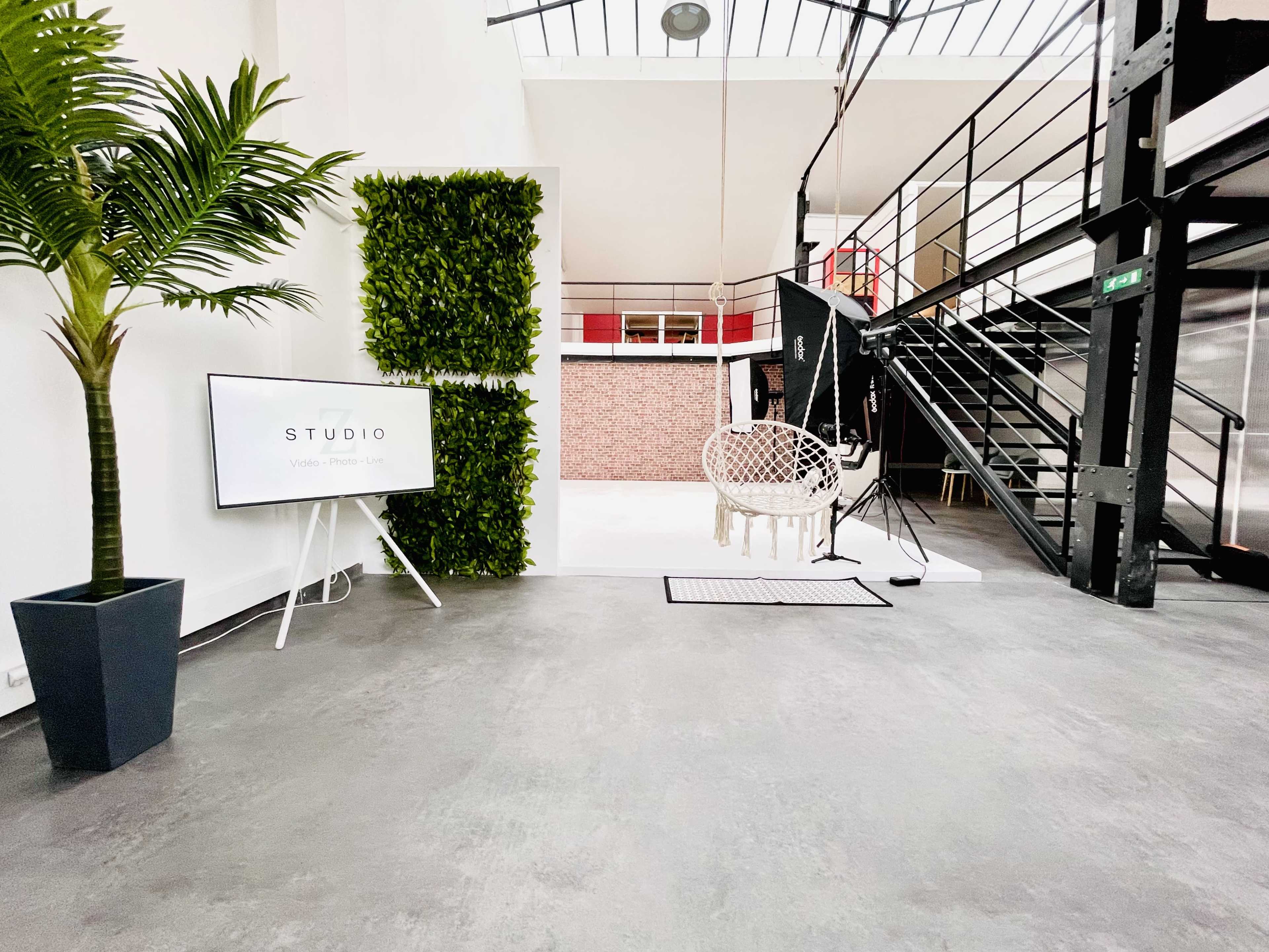A modern studio space featuring a hanging swing chair, a large screen displaying "STUDIO," and a green wall, with industrial-style stairs in the background.