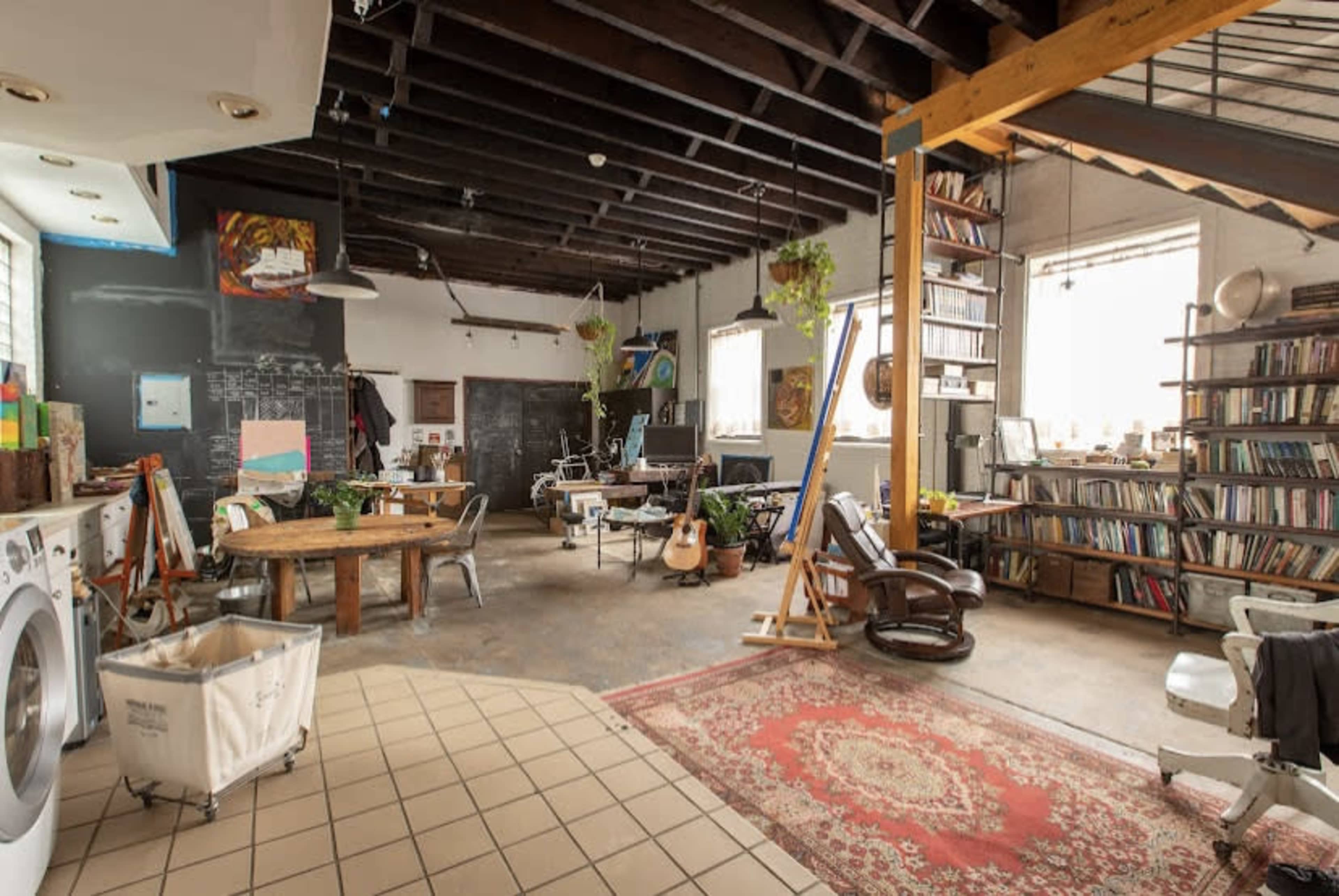 A spacious, open studio featuring wooden beams, a mix of furniture, bookshelves filled with books, and various art supplies scattered throughout.