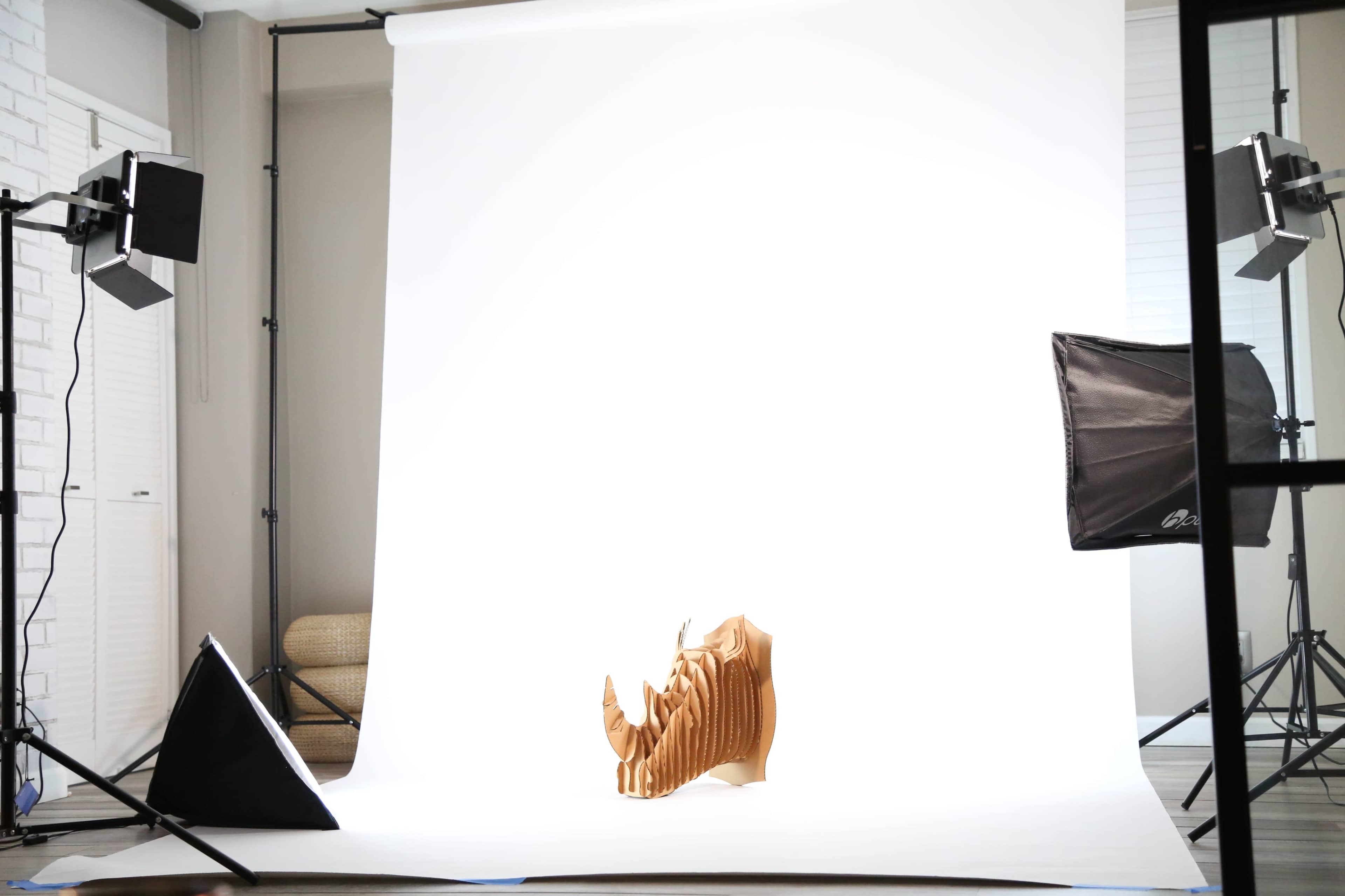 The image shows a wooden sculpture of a rhinoceros positioned against a bright white backdrop in a well-lit studio setting.