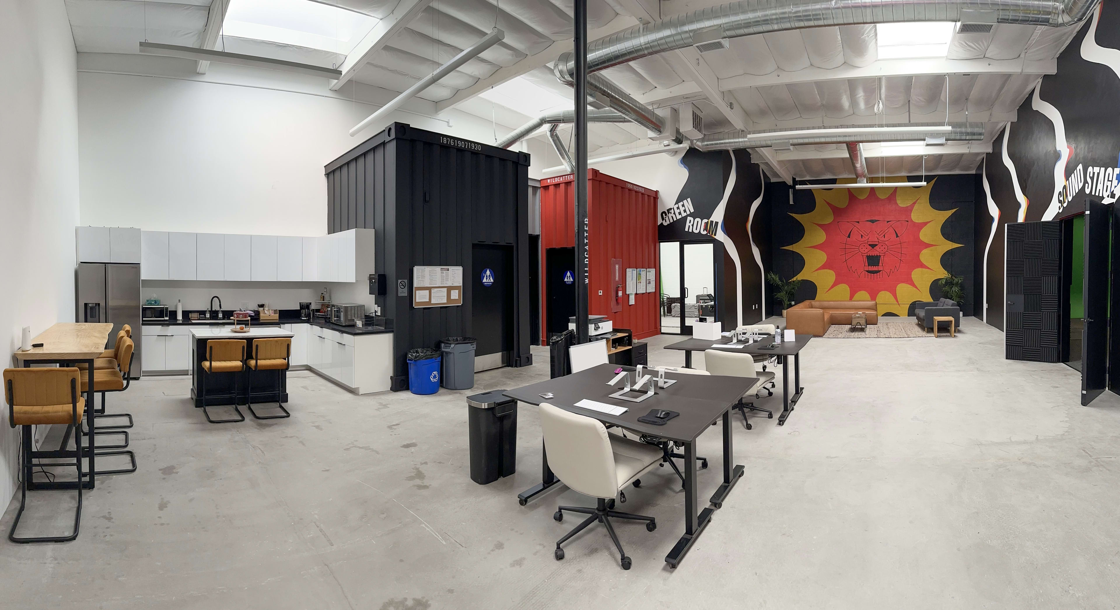 A spacious, modern office interior featuring a kitchen area, workstations, and a vibrant mural on the wall.
