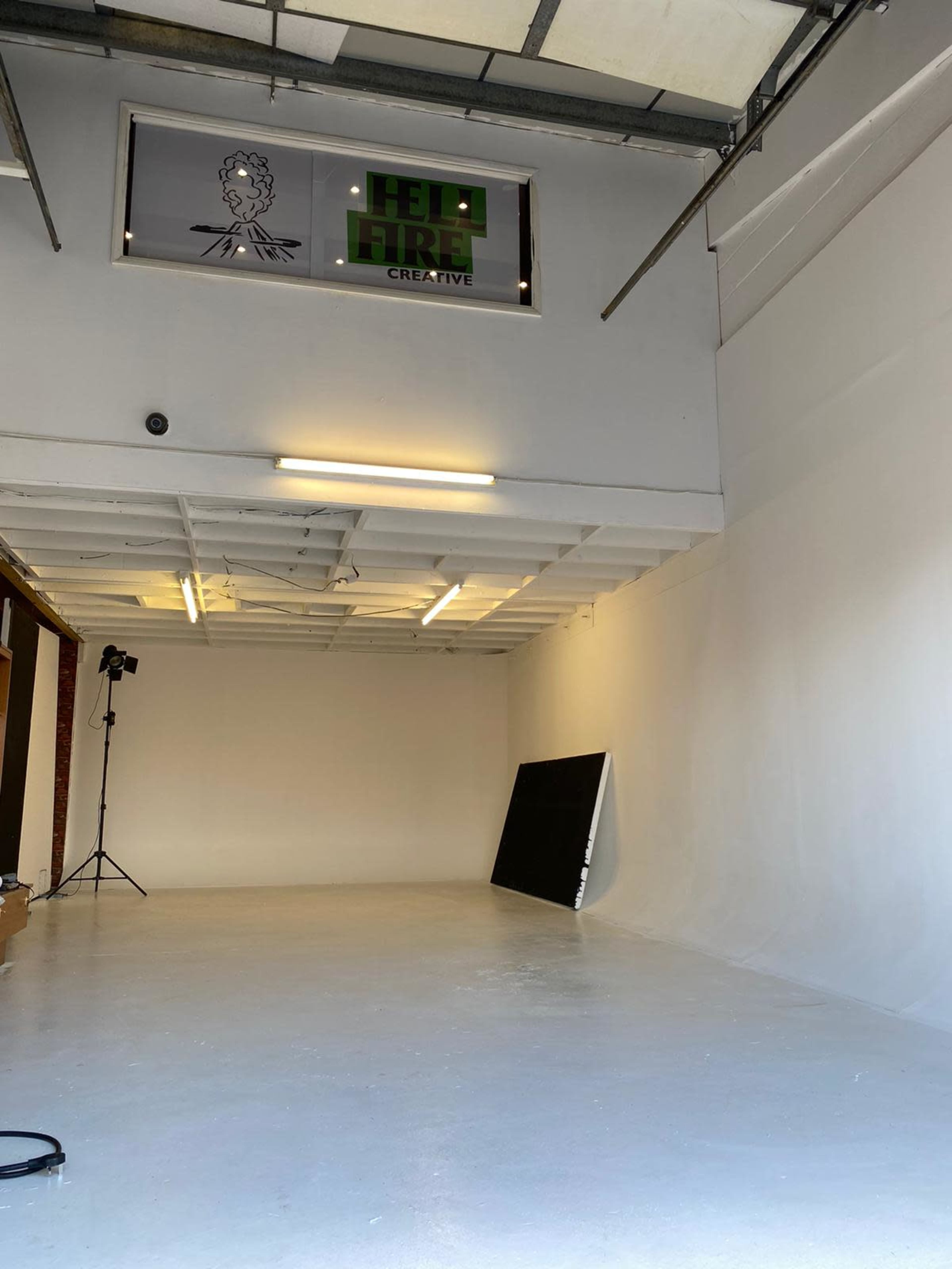 A spacious, empty studio with a white wall, a large black board leaning against it, and a sign for "HELP HIRE CREATIVE" on the wall above.