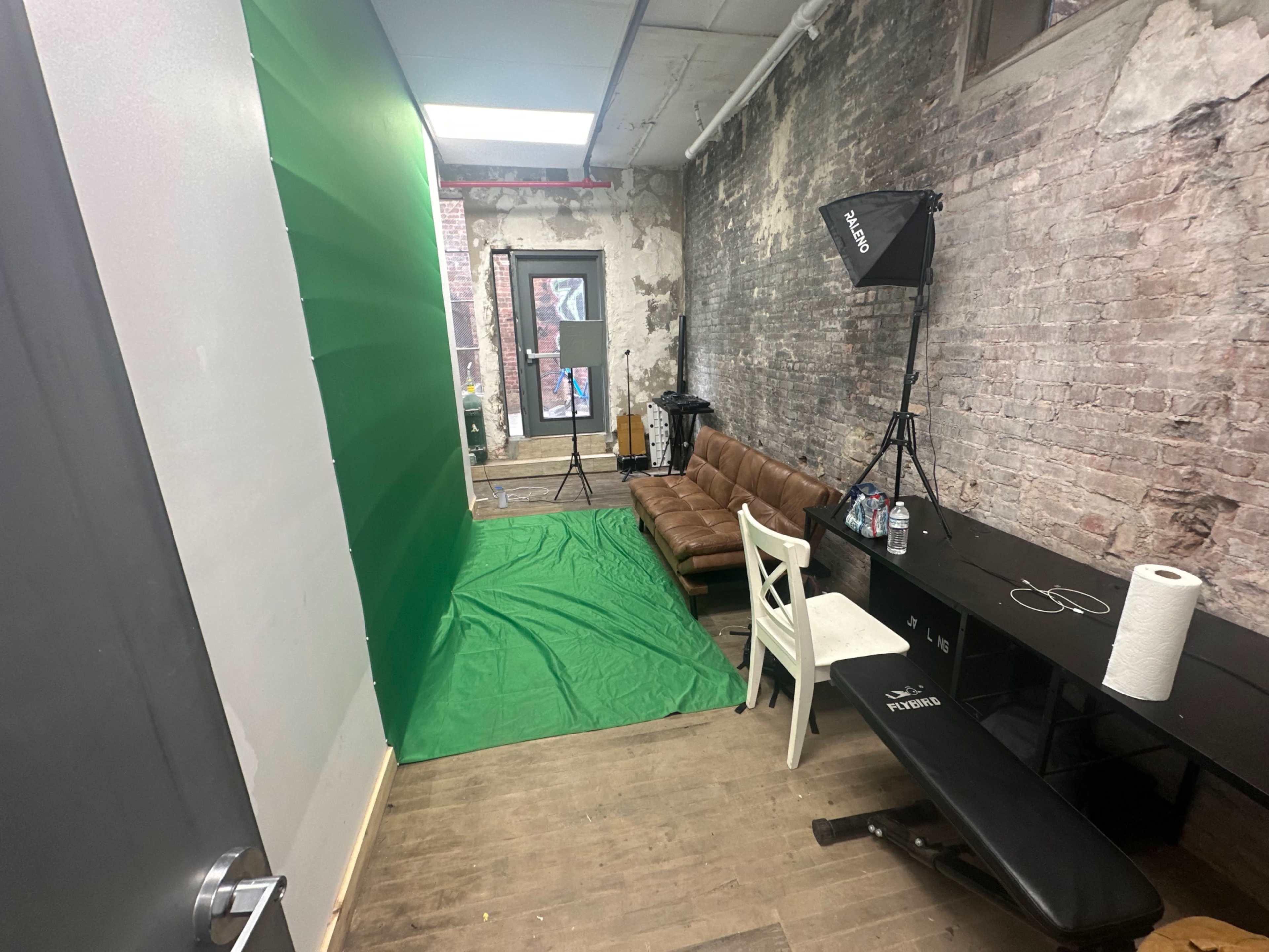 The image shows a small room with a green screen setup, a brown couch, a desk with a roll of paper towels, and studio lights positioned nearby.