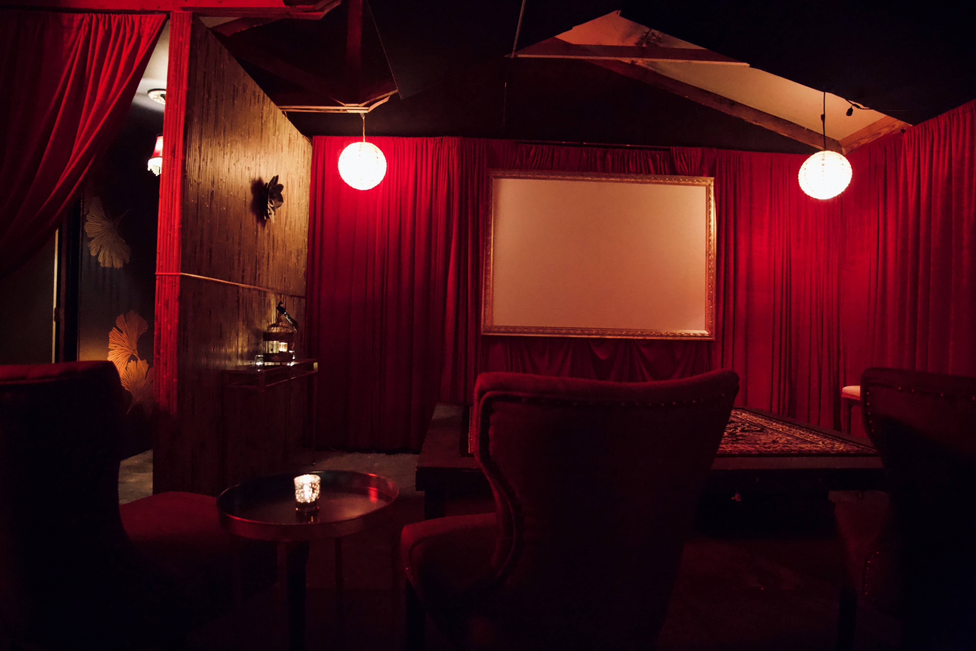 A dimly lit room featuring red curtains, a blank screen, and plush seating arranged around a small table.