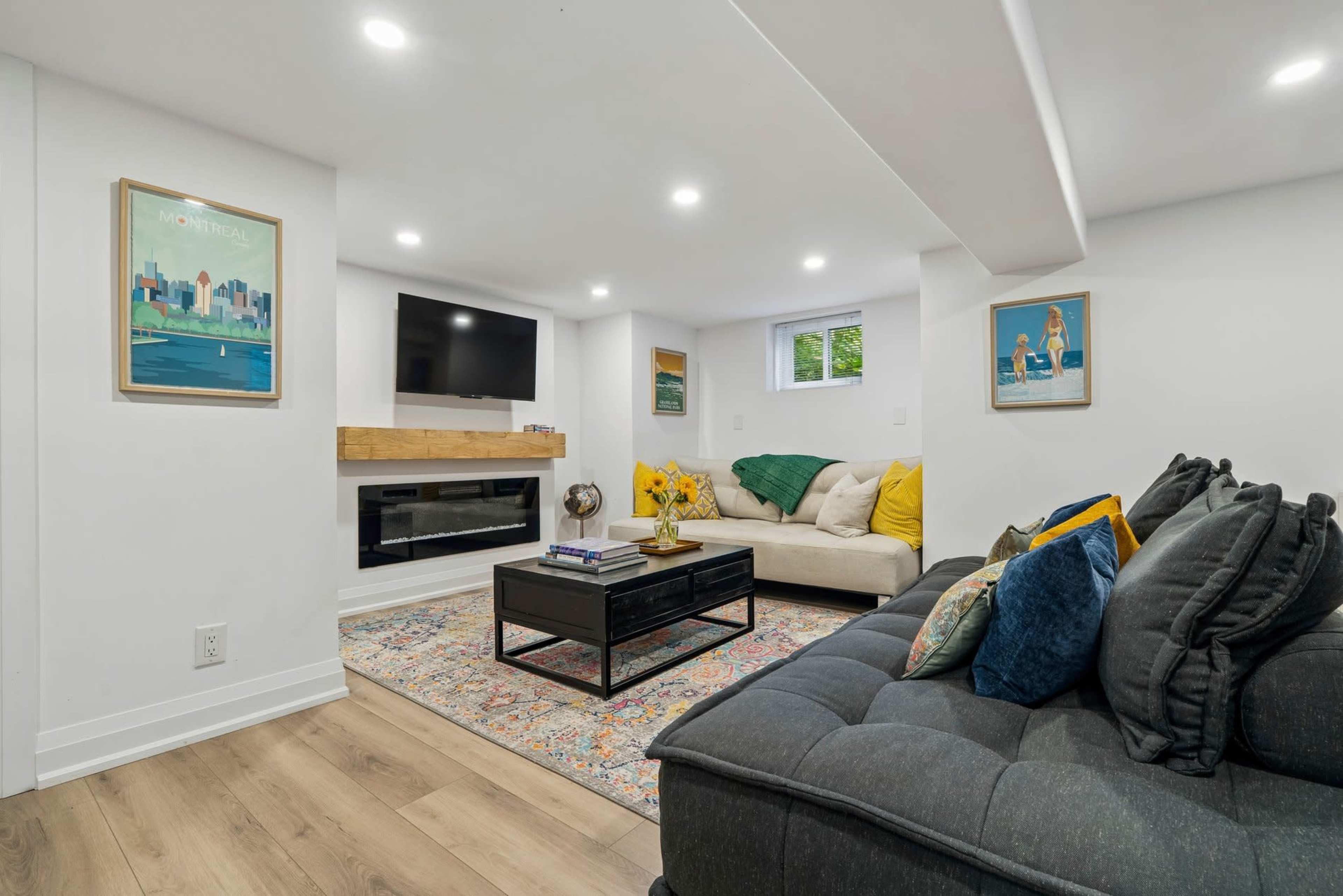 The image shows a modern basement living room with a television mounted on the wall, a sofa, a coffee table, and a fireplace.