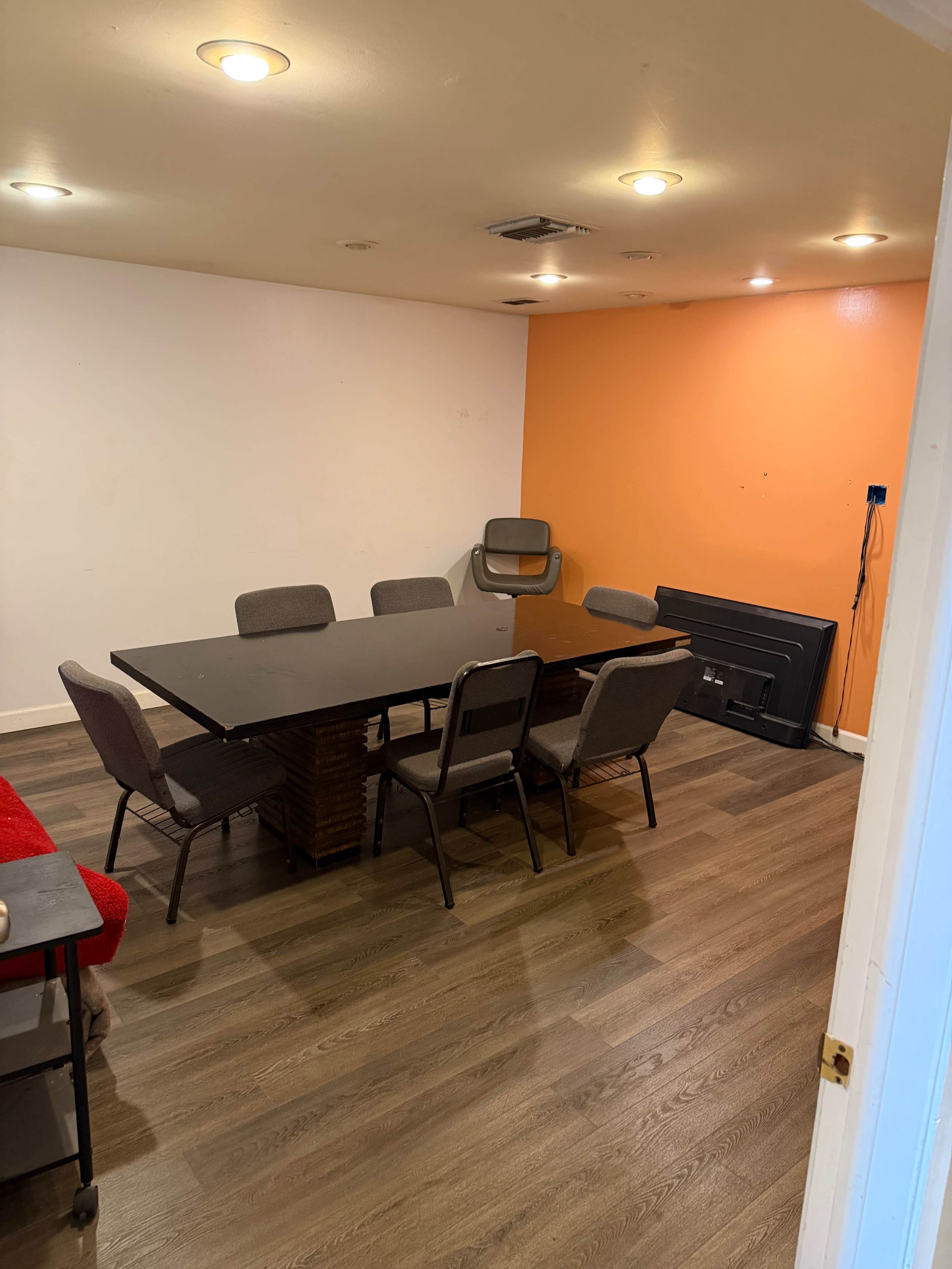 The image shows a room with a large rectangular table surrounded by several chairs, accented by one orange wall and a floor made of laminate wood.