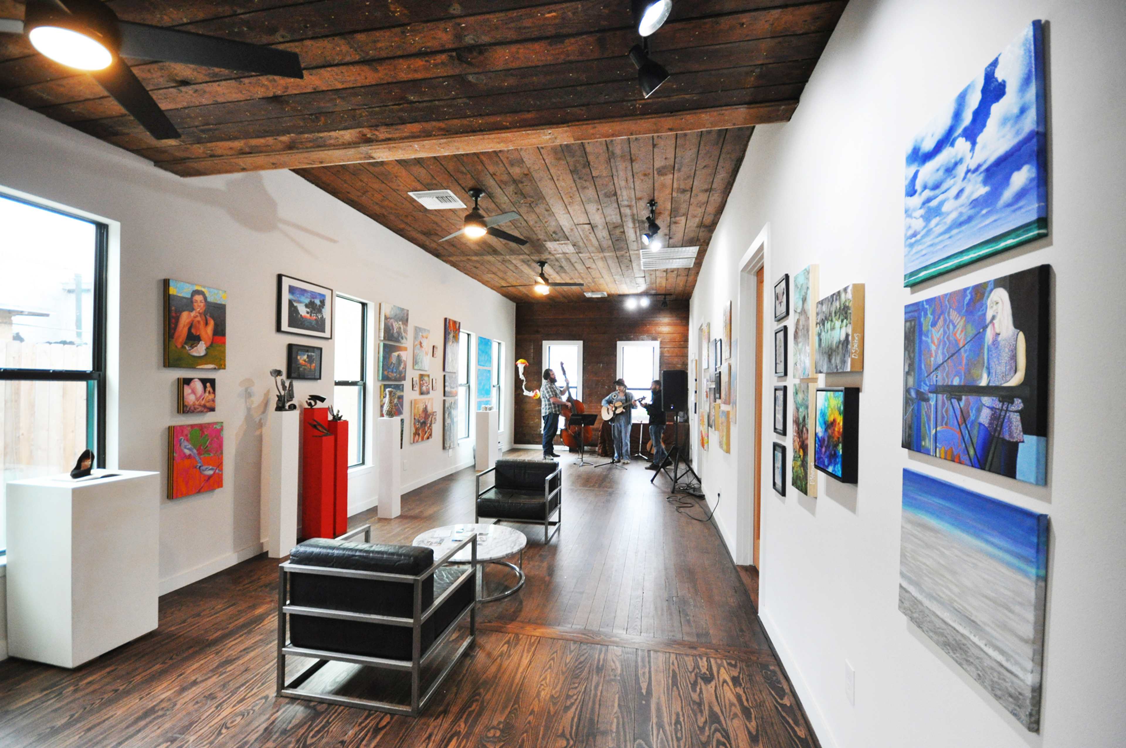 An art gallery with wooden floors and a variety of colorful paintings displayed on the walls, while a musician performs in the background.