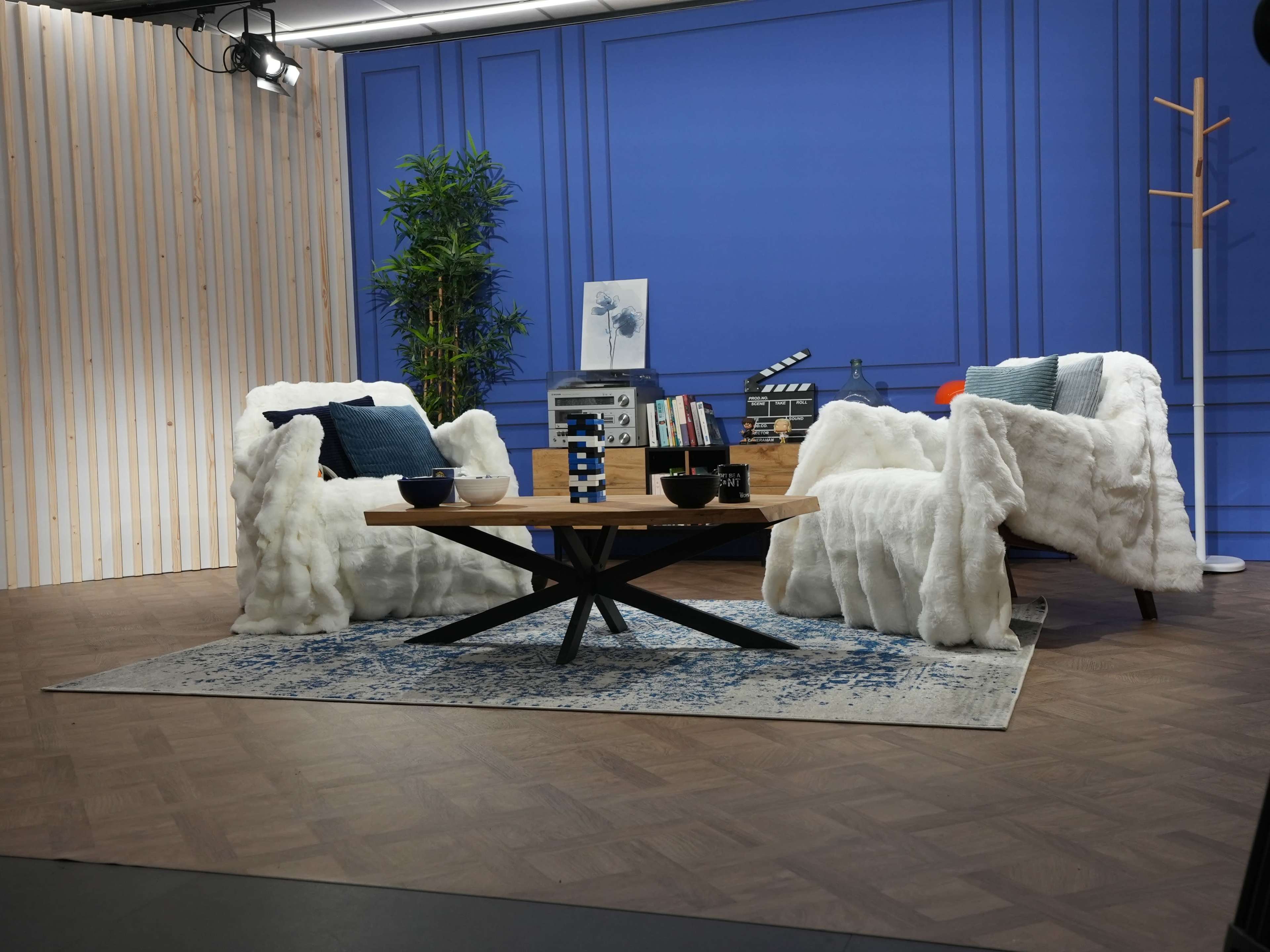 The scene features two white chairs draped with cozy throws, a round wooden coffee table, and a blue backdrop, creating a modern lounge setting.