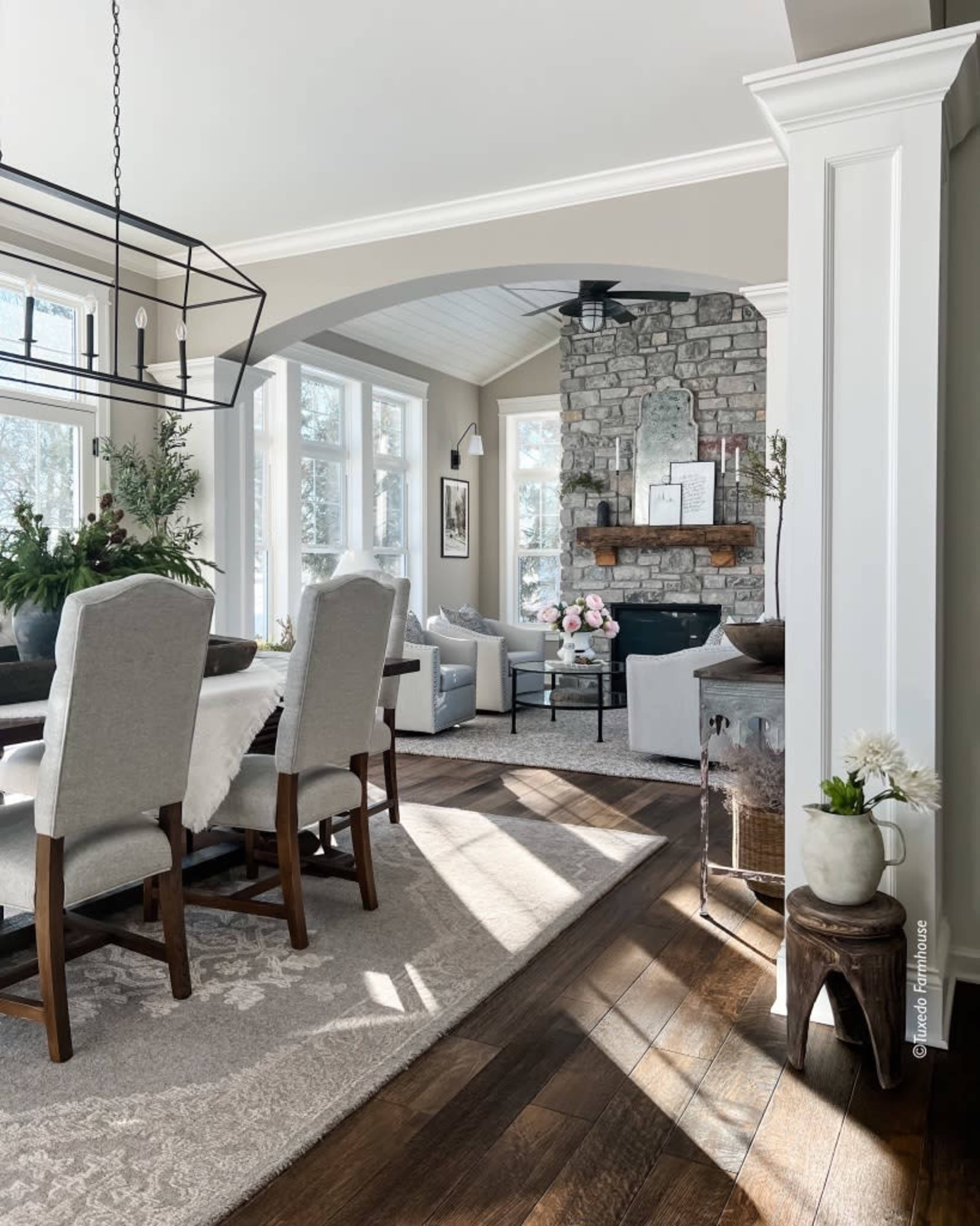 A bright, inviting living space featuring a dining area with upholstered chairs, a stone fireplace, and large windows allowing natural light to fill the room.